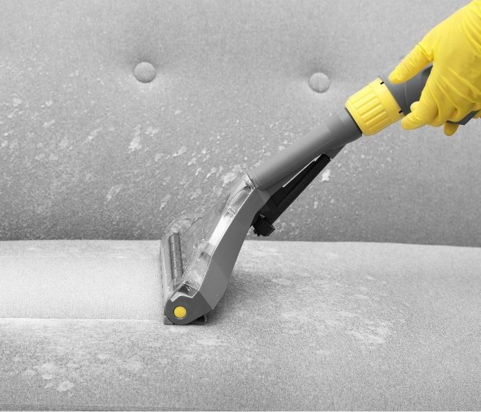 A person wearing yellow gloves is cleaning a couch with a vacuum cleaner