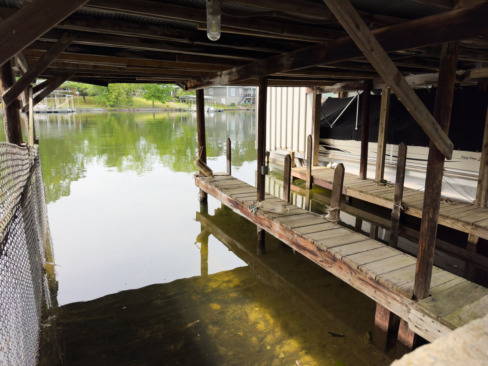 Wooden dock under a boathouse over calm water, with bright green trees reflected nearby.