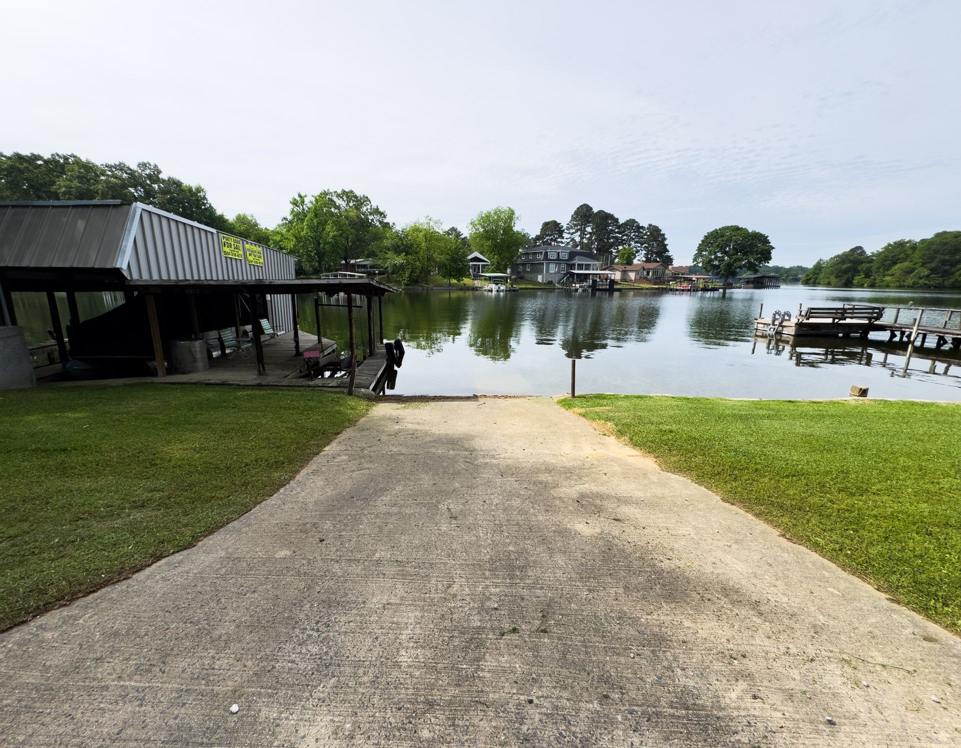 Paved path leading to a calm lake with a dock, covered pavilion, and trees along the shore
