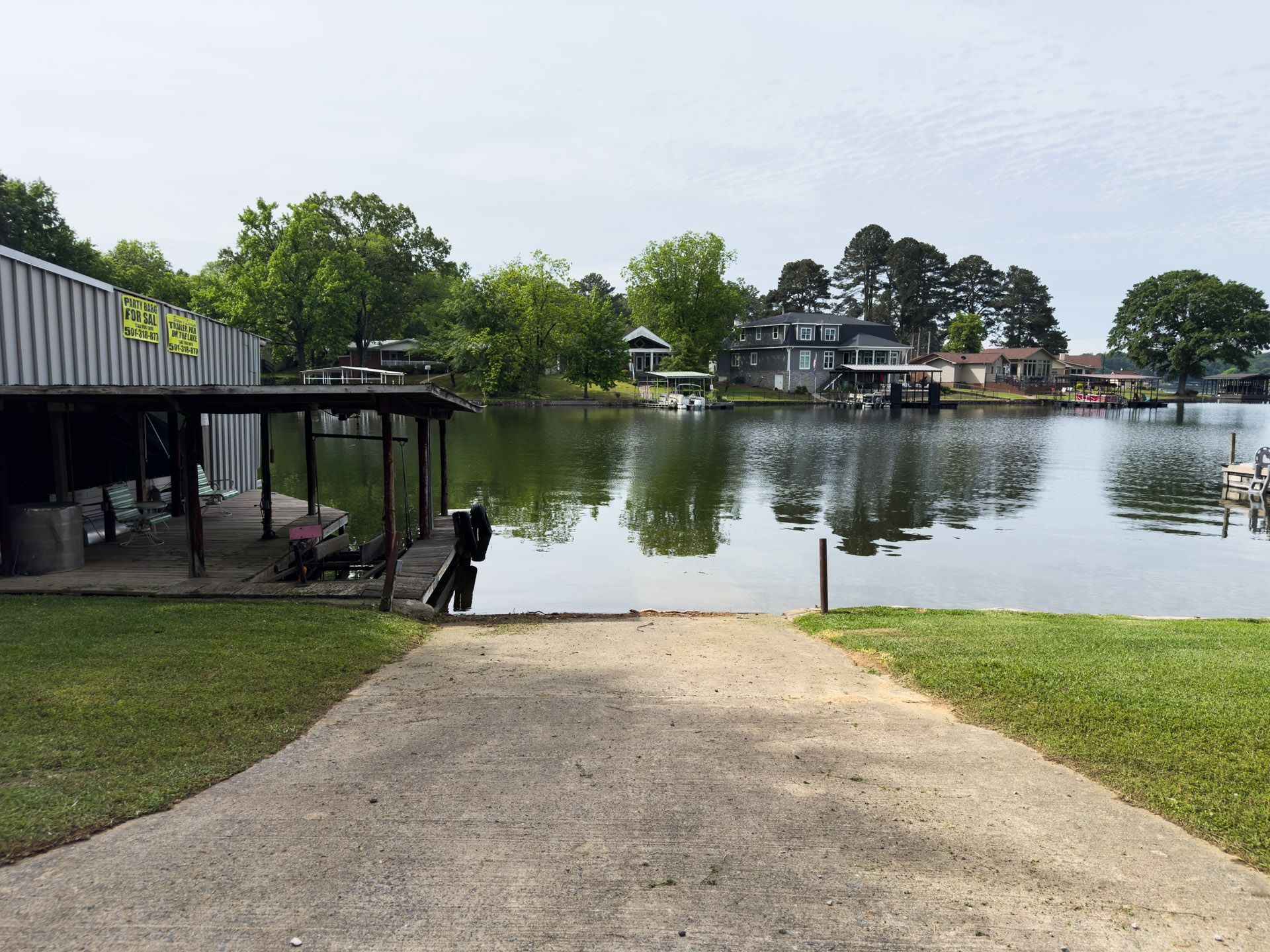 Gravel boat ramp leading to a calm lake with dock, trees, and houses on the far shore