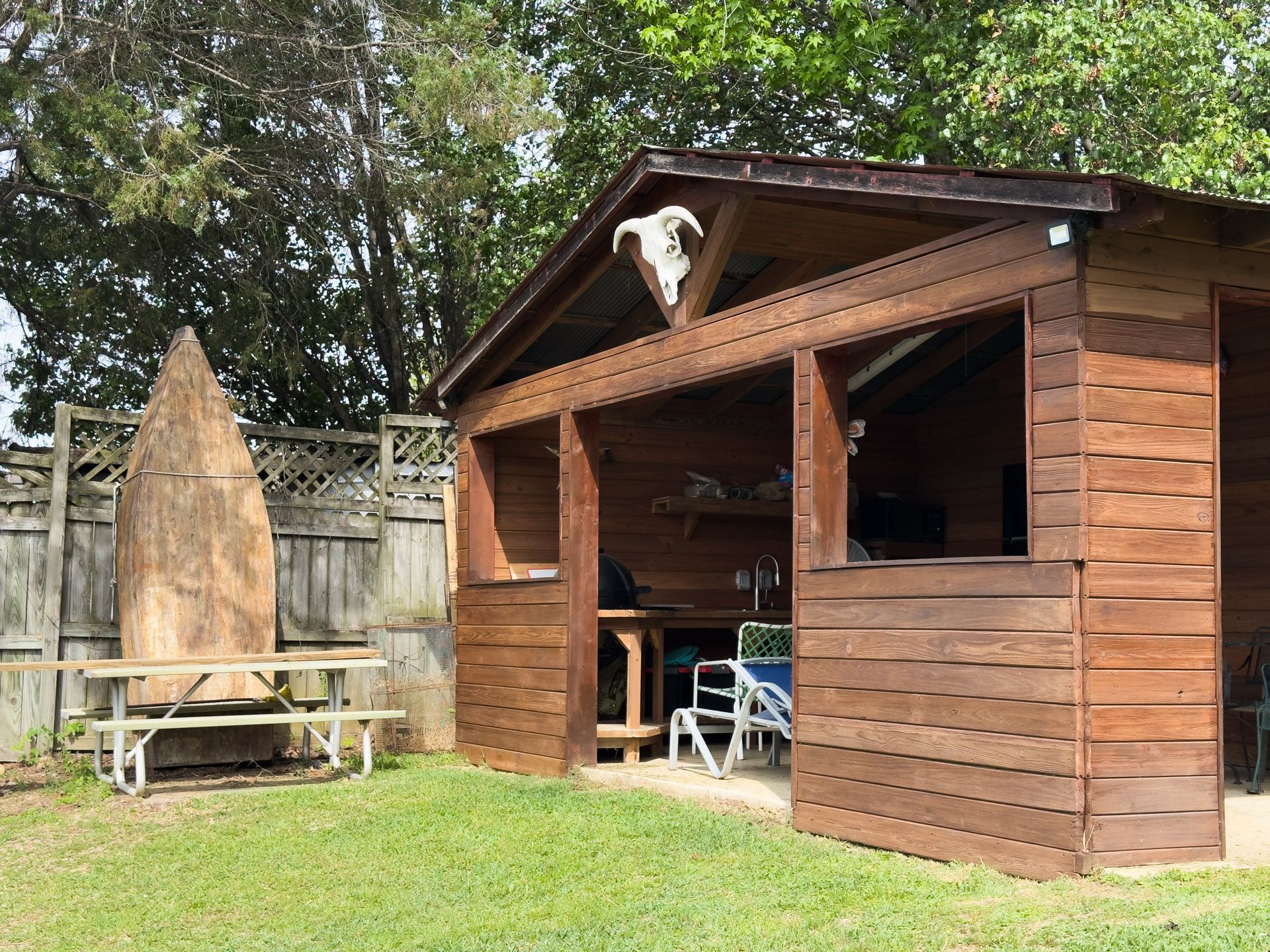 Wooden garden shed with open door, picnic table, and surfboard leaning by a fenced backyard.