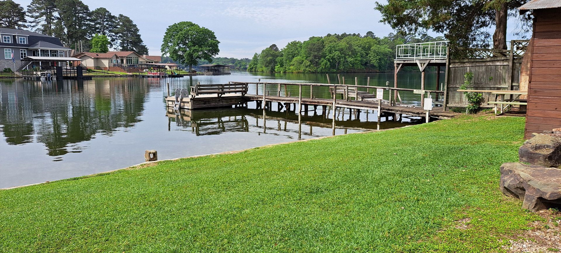 Grassy lakeside with wooden docks and boats, calm water, trees, and houses along the shore