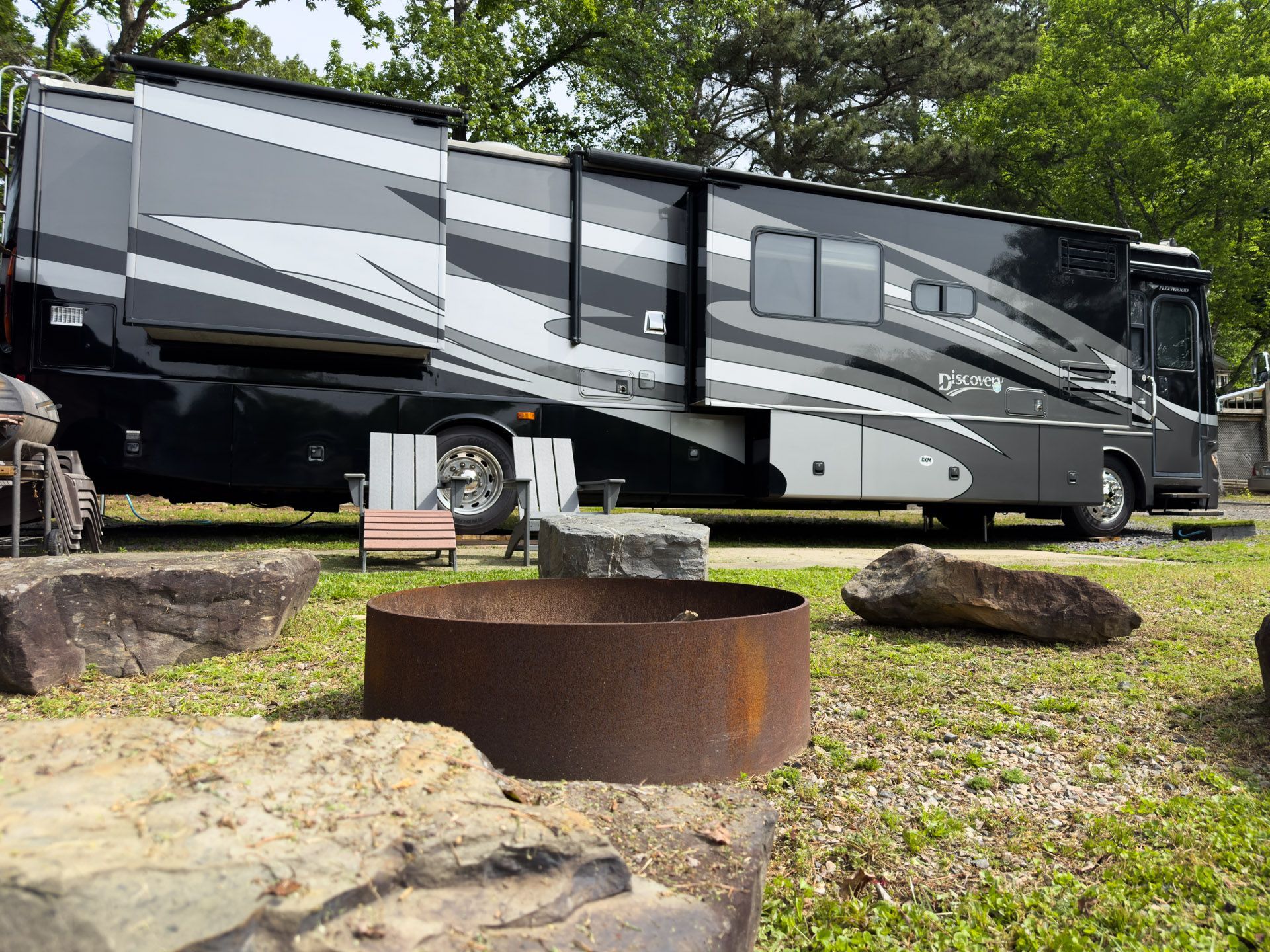 Large gray motorhome parked at a wooded campsite beside picnic tables and fire rings.