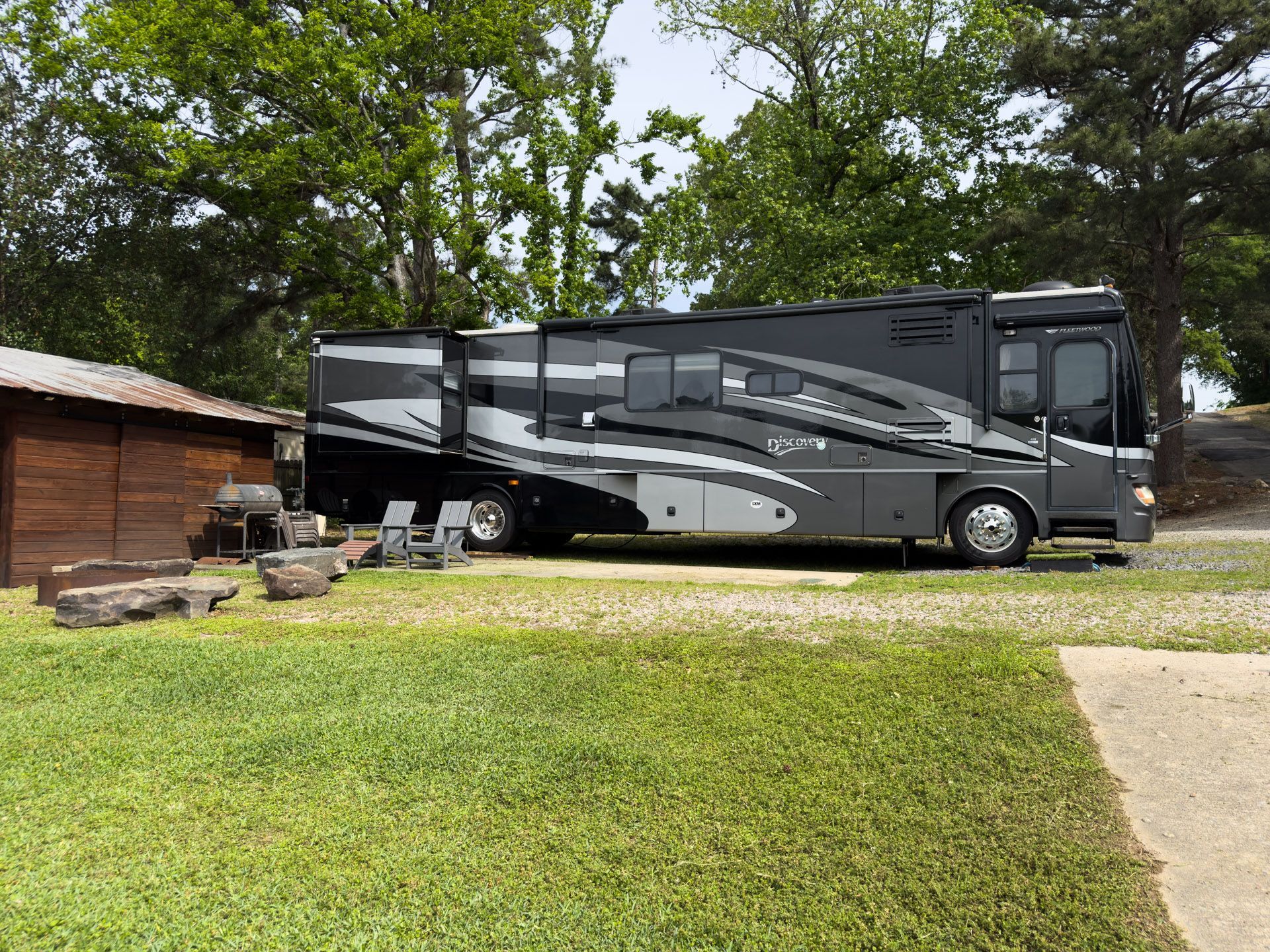 Large black-and-gray RV parked on grass beside a small wooden cabin and trees
