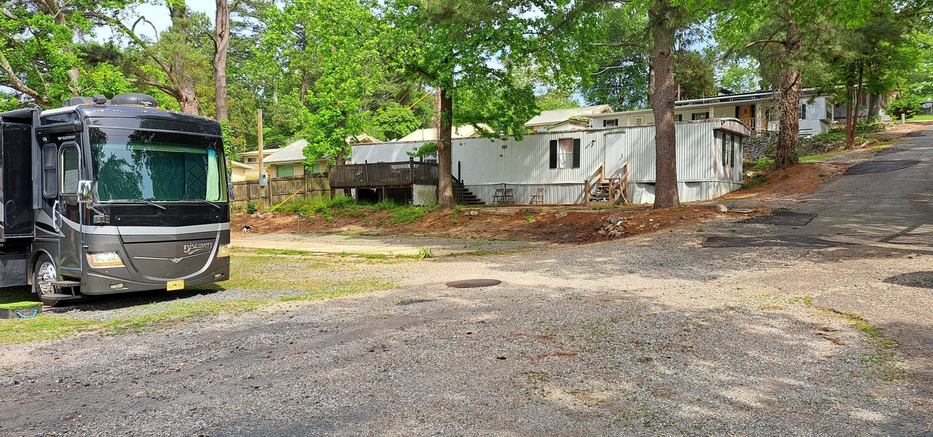 Large RV parked beside small white mobile homes on a gravel lot in a wooded area