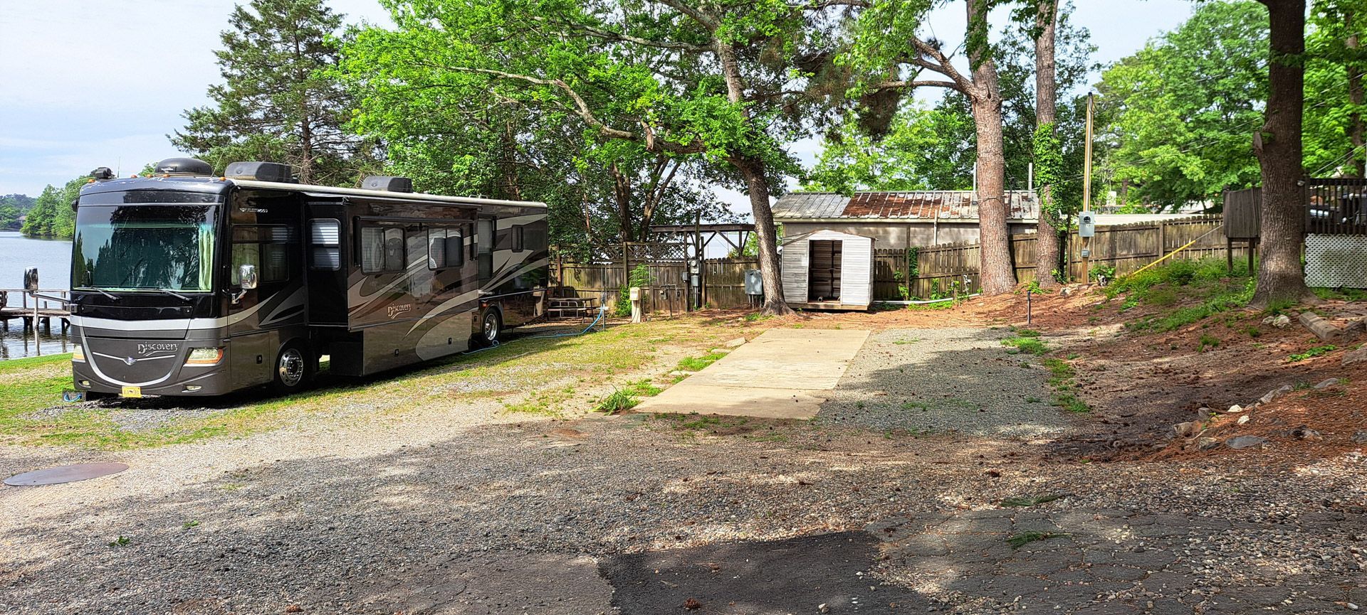 Black tour bus parked on a gravel lot beside a small building and trees on a sunny day