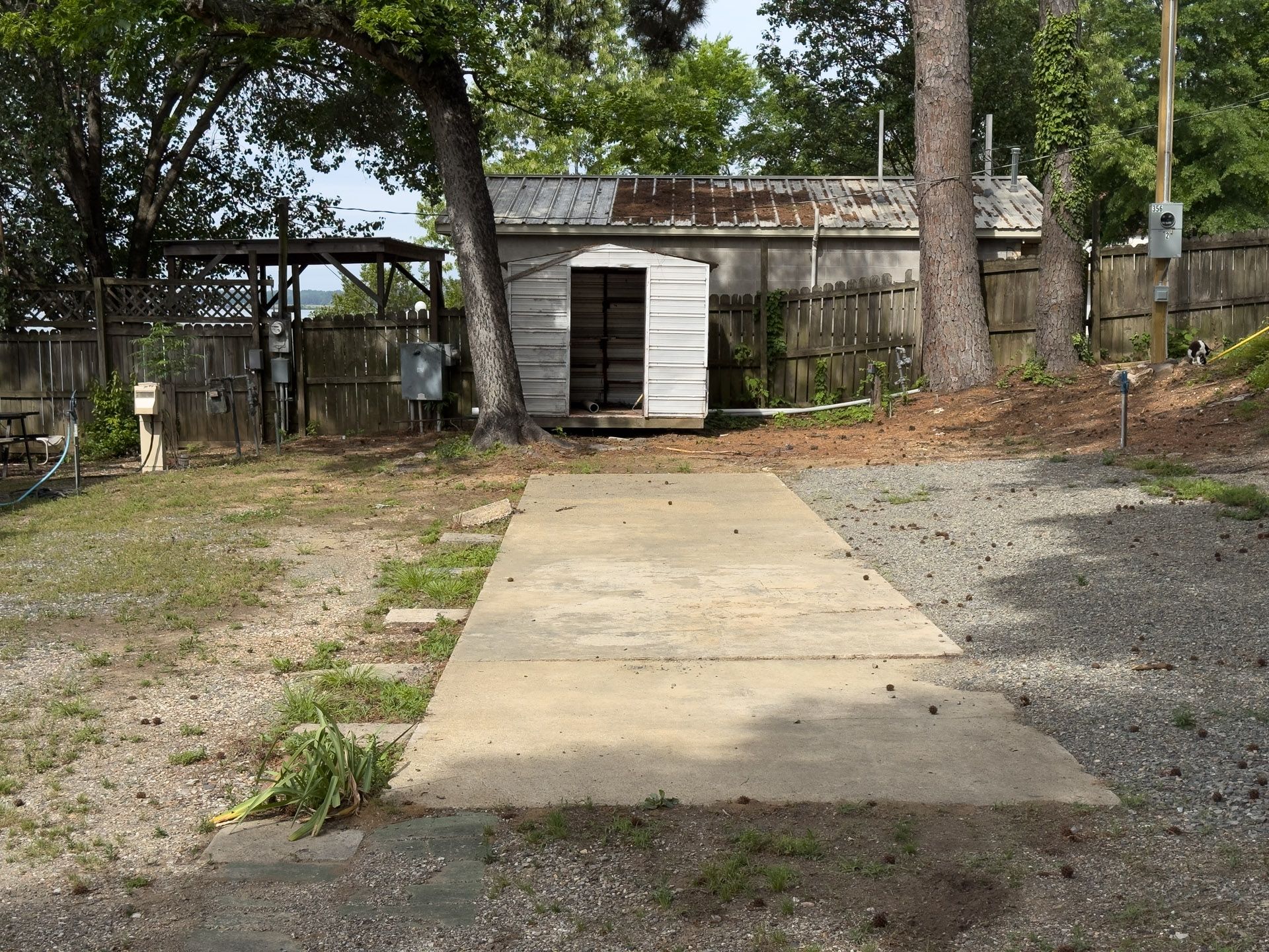 Concrete driveway leading to a small white shed in a wooded backyard.