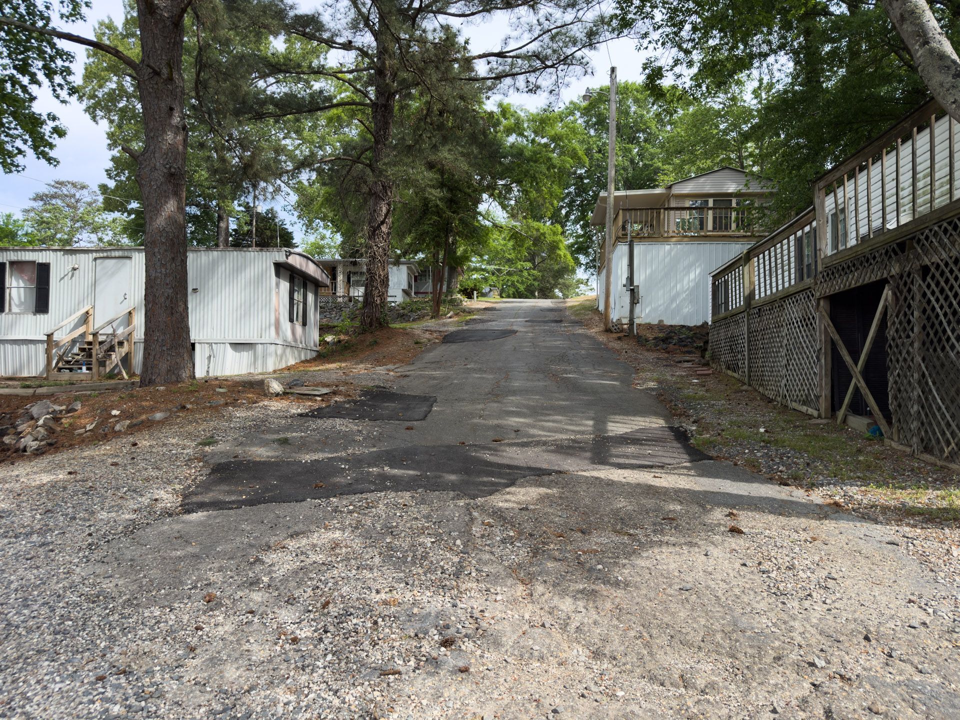 Gravel road lined with trees and small buildings, leading uphill beside a white fence and wooded area.