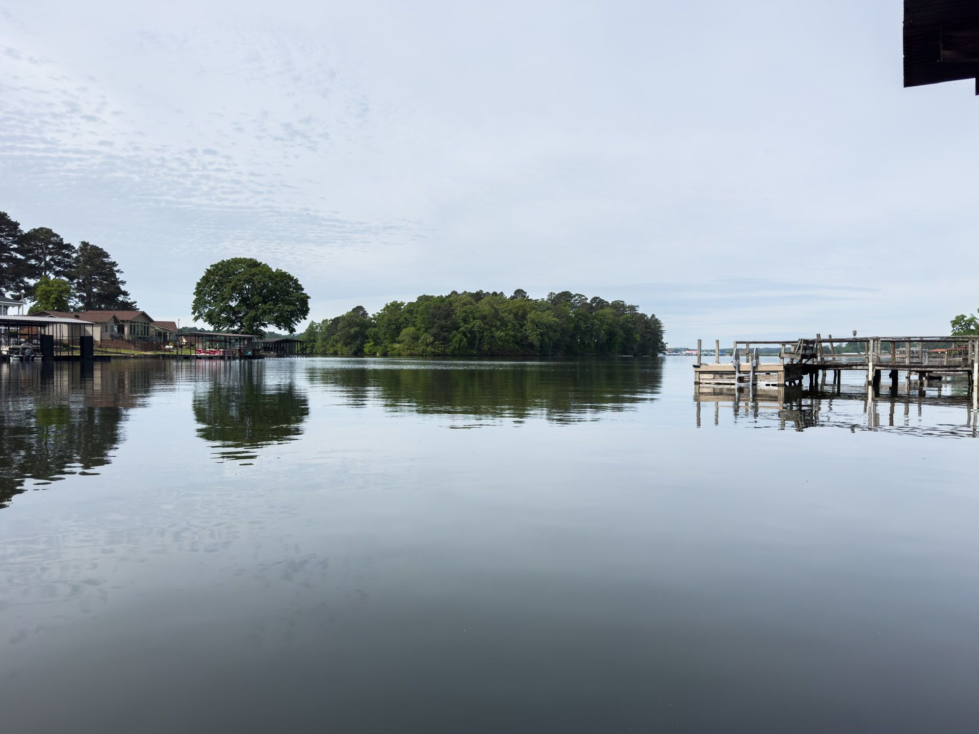 Calm lake with tree-lined shore, wooden docks, and buildings reflected on still water under a cloudy sky