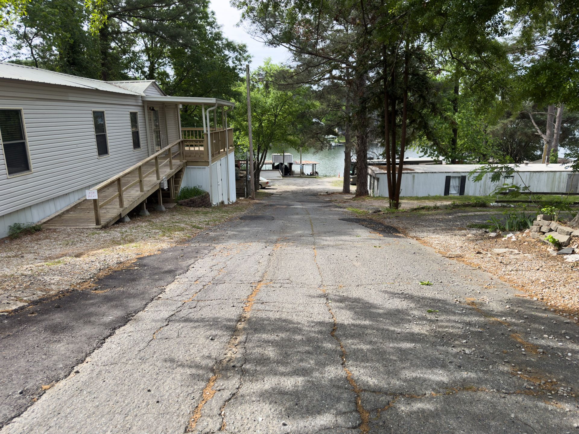 Gravel driveway beside a white mobile home, leading downhill toward trees and a lake.