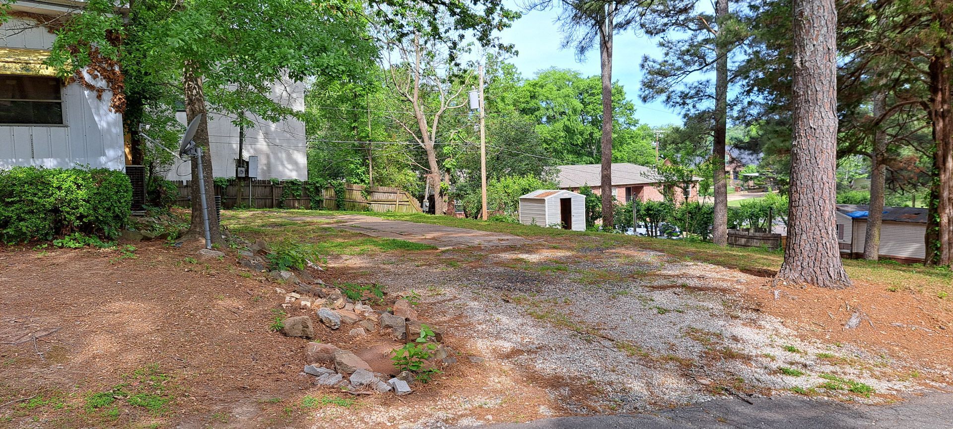 Wooded yard with pine trees, gravel driveway, and small houses in the background