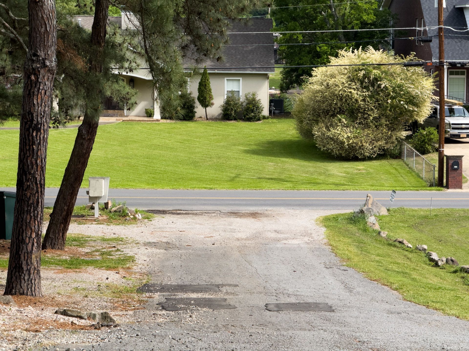 Gravel driveway leading to a suburban street with green lawn, trees, and houses in the background