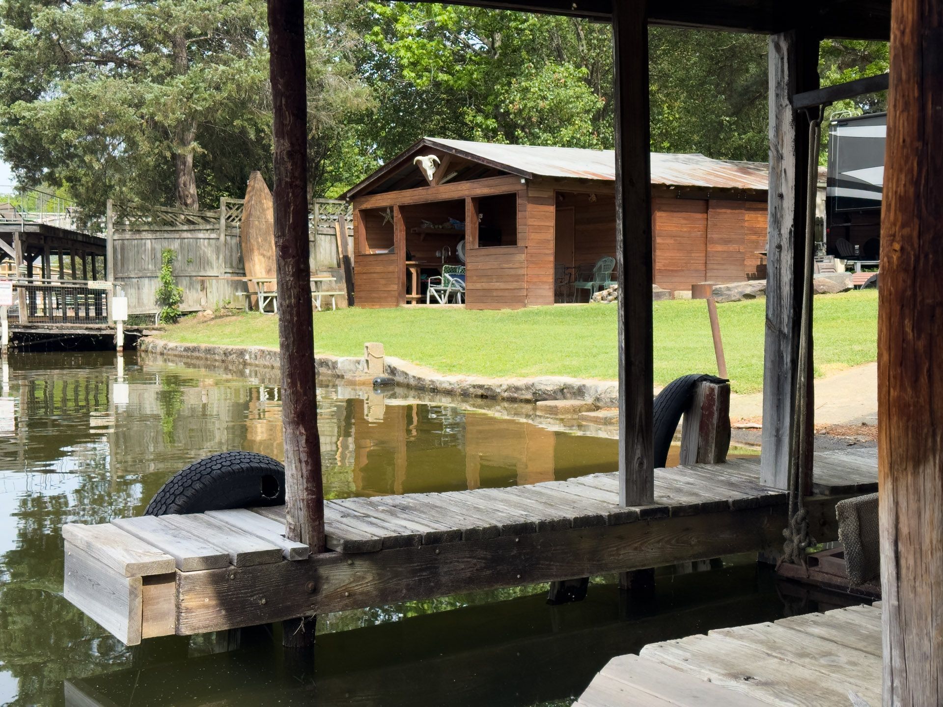 Wooden dock beside a calm pond, with a small brick cabin and grassy yard in the background.
