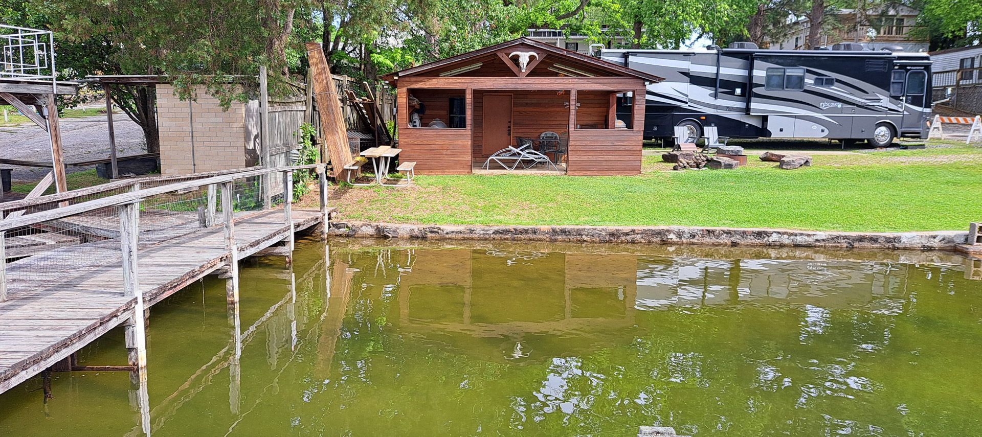 Wooden dock beside a green pond, with a small cabin and parked cars in the background