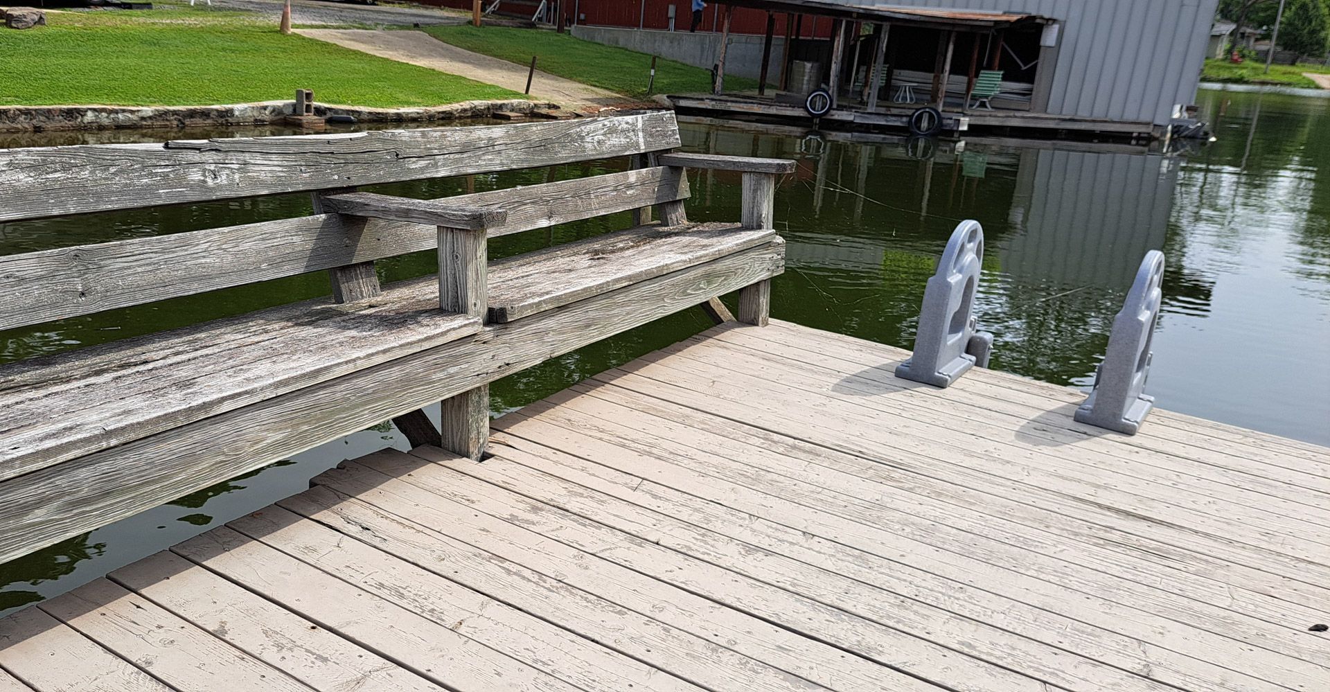 Weathered wooden dock beside calm water, with a bench and covered boat slip in the background