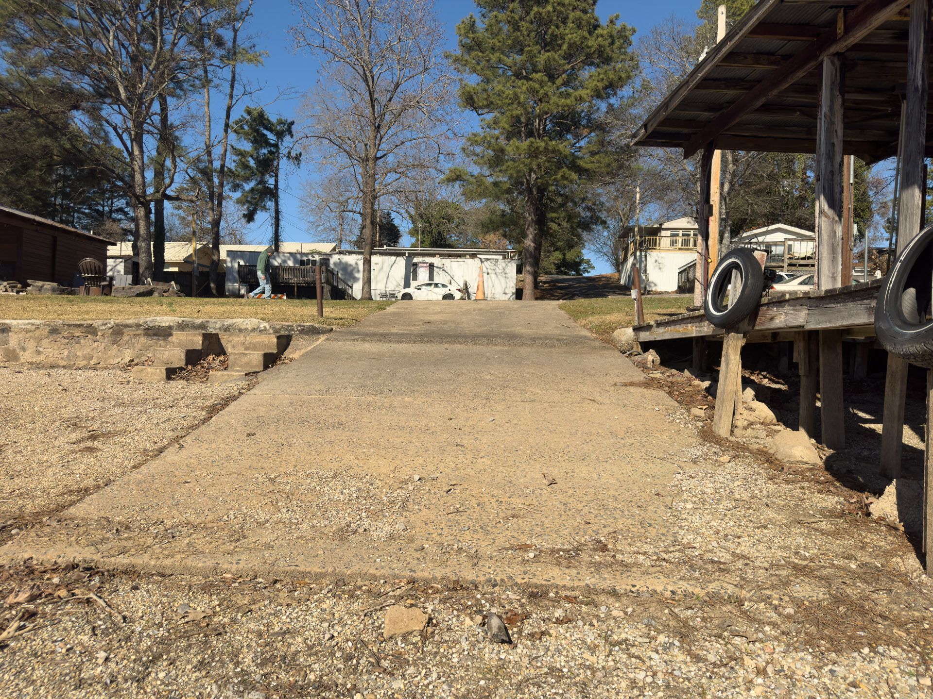 Concrete boat ramp leading toward mobile homes. Wooden dock with tires on the right. Gravel shoreline.