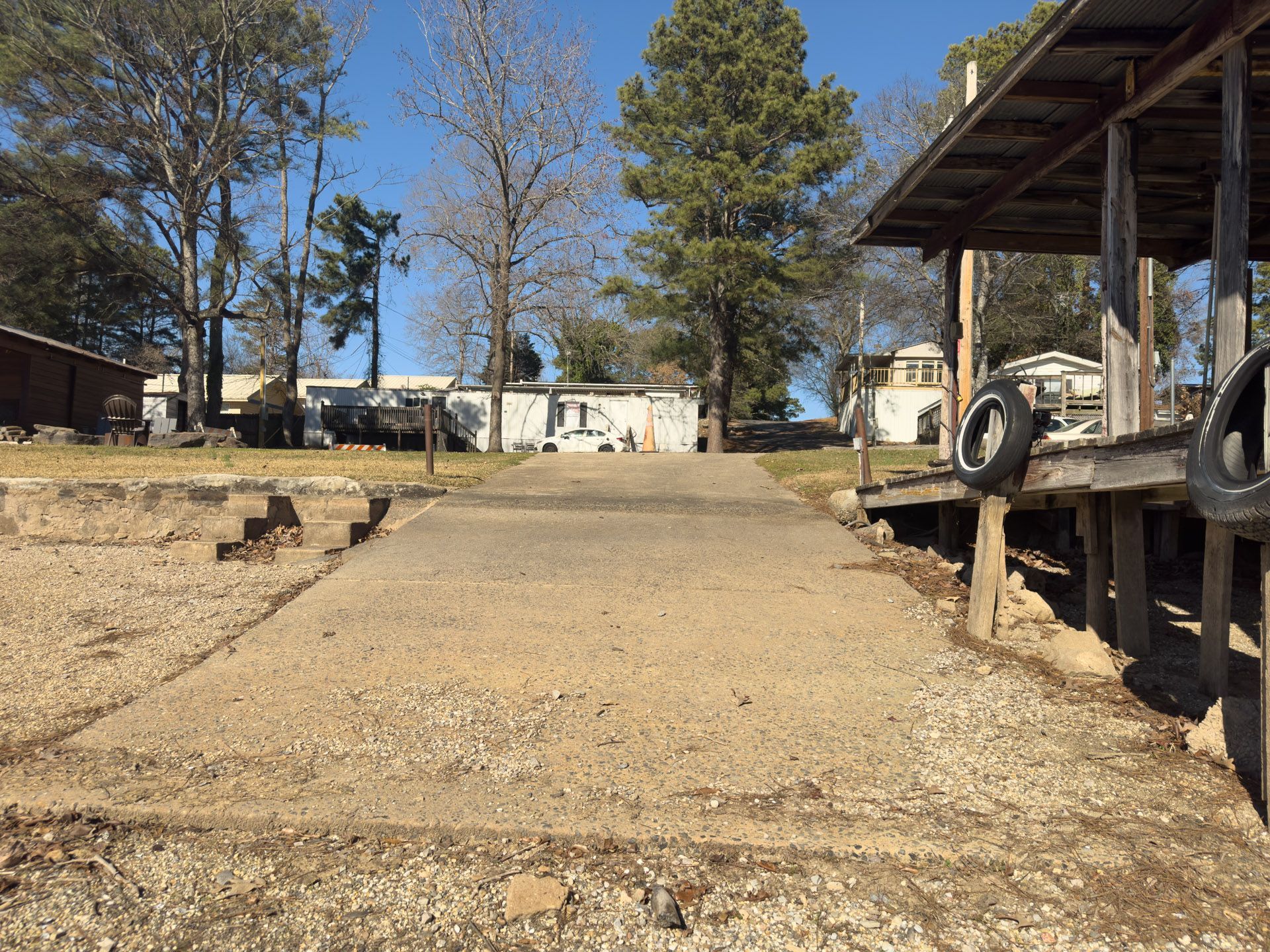 Gravel boat ramp leads to a covered dock, trailers and trees in background, clear sky.