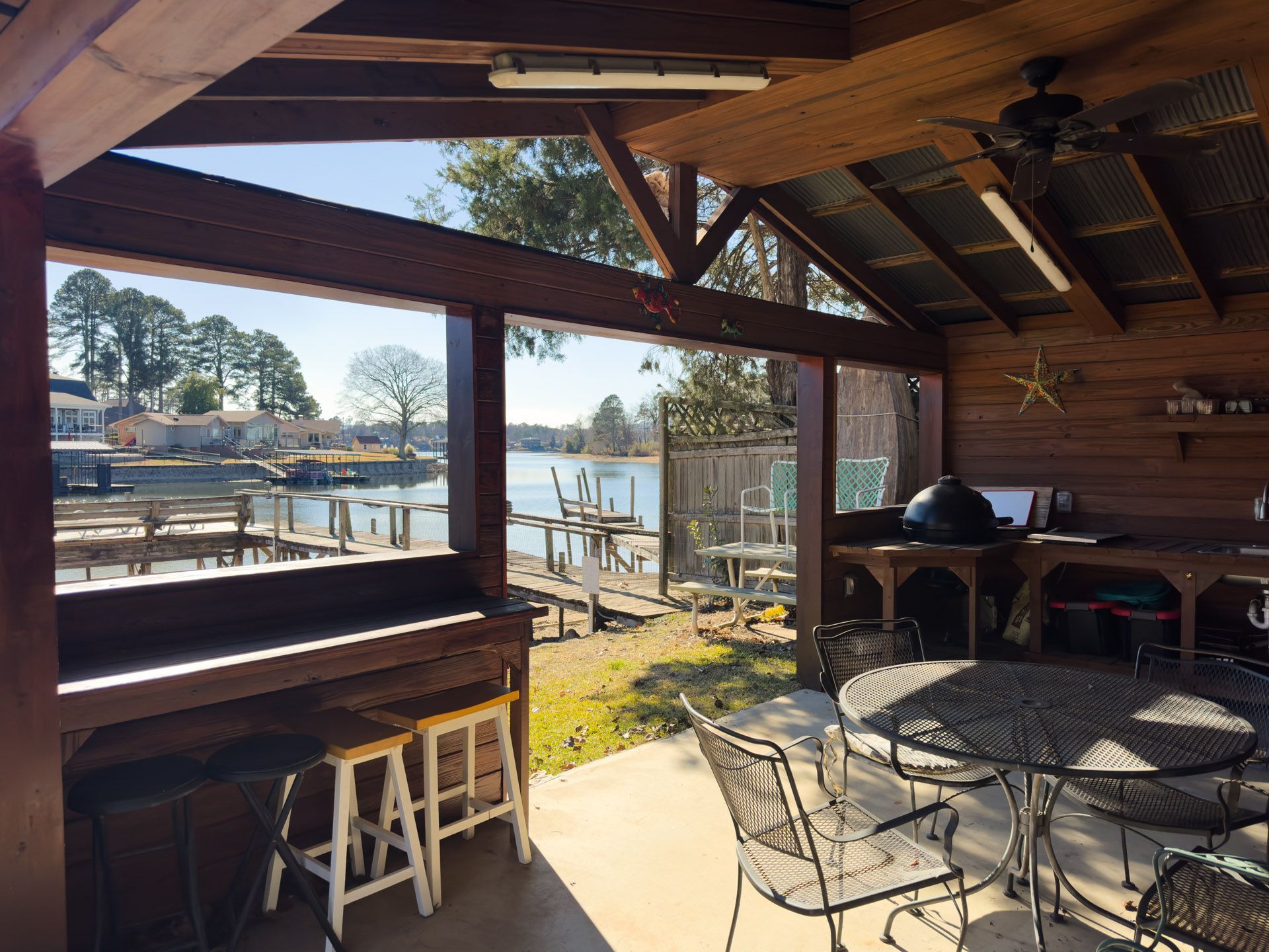 Covered patio overlooking water. Dark wood frame, metal furniture, grill, dock, trees, and blue sky.