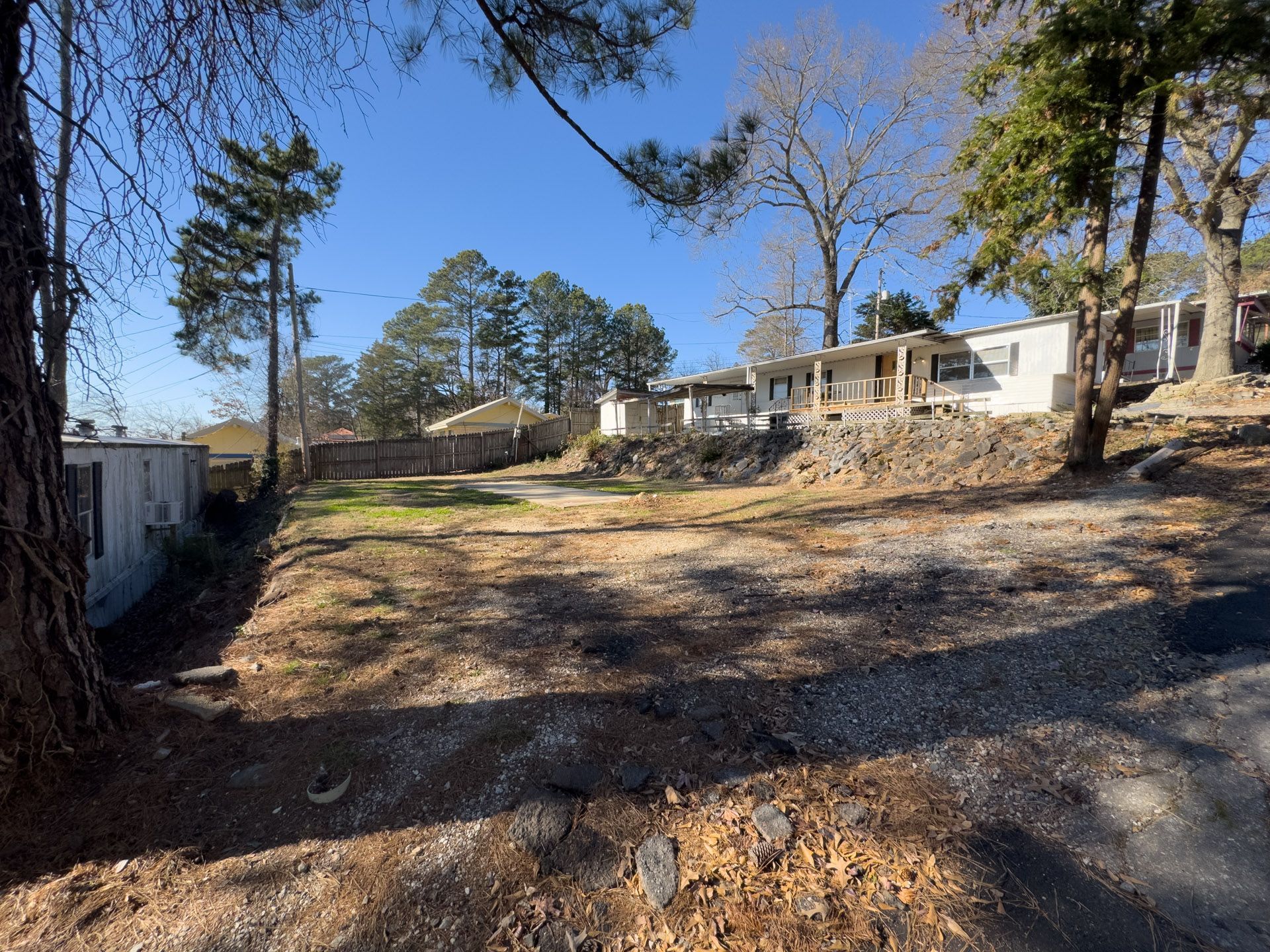 Cleared lot with dirt and grass, small houses on a hill, trees, and a clear blue sky.