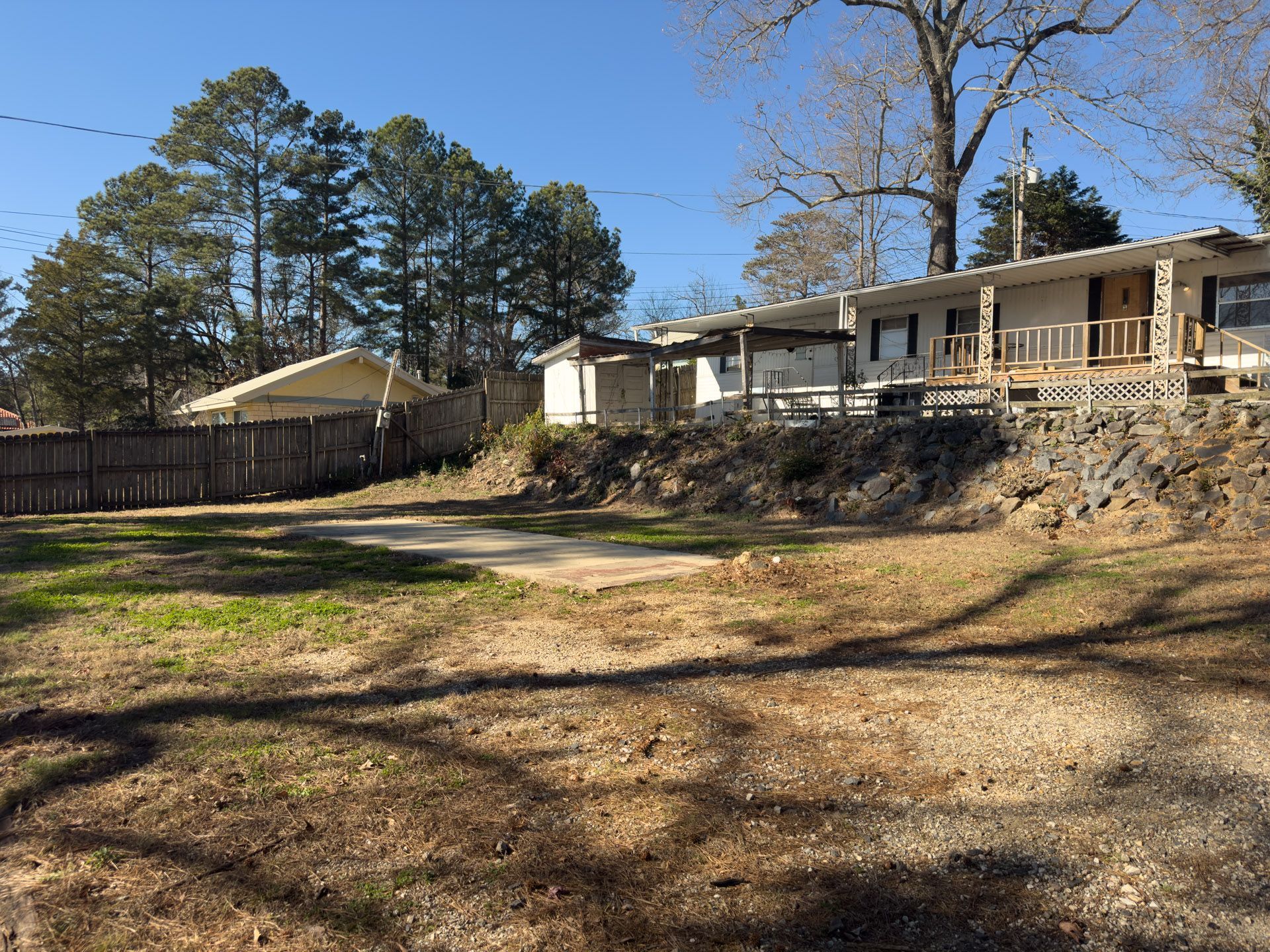 Brown, grassy lot with a house on a rocky hill. Wooden fence and trees in background. Bright, sunny day.