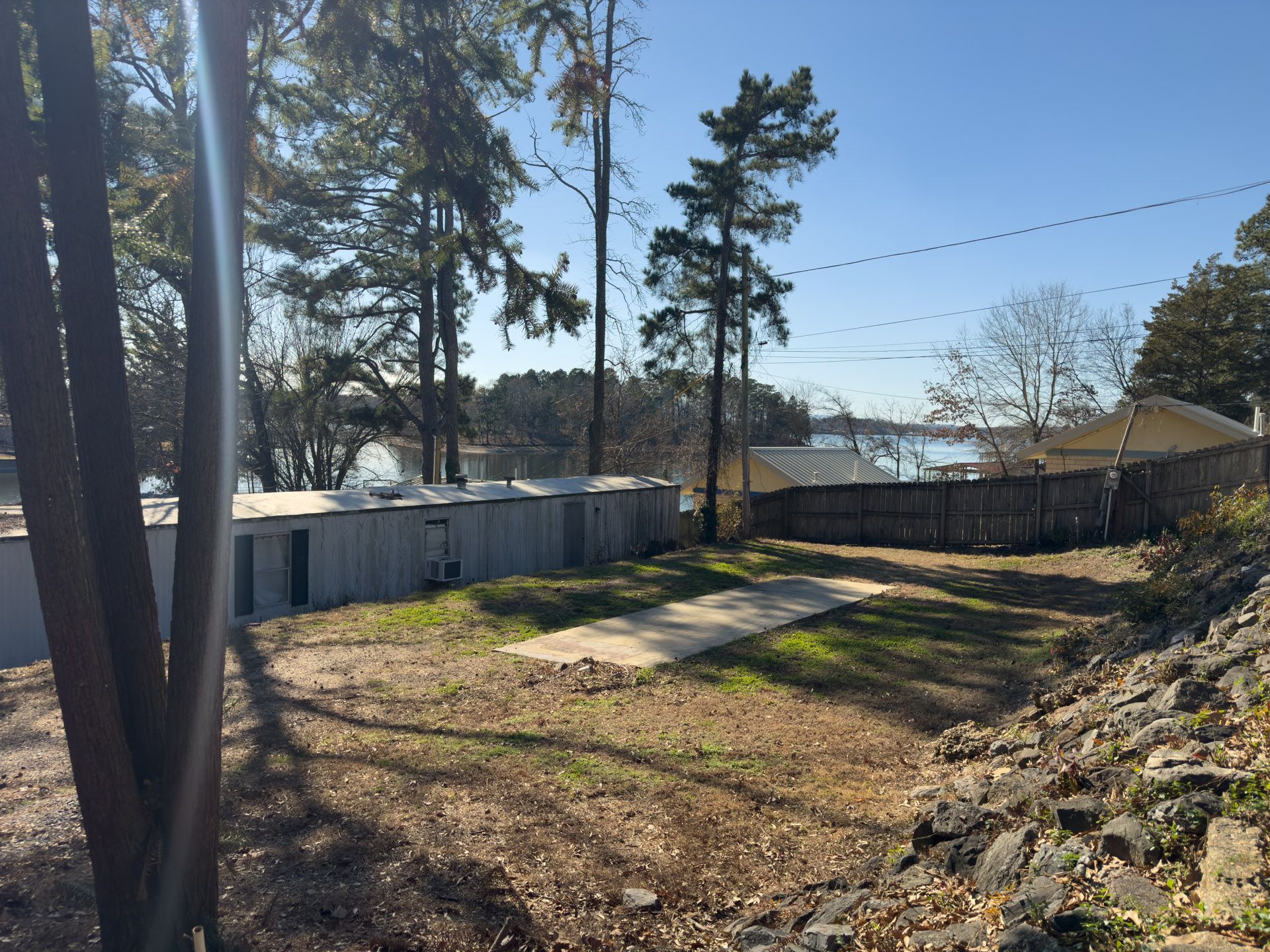 View of a grassy area, concrete pad, and buildings near a body of water with trees and a blue sky.