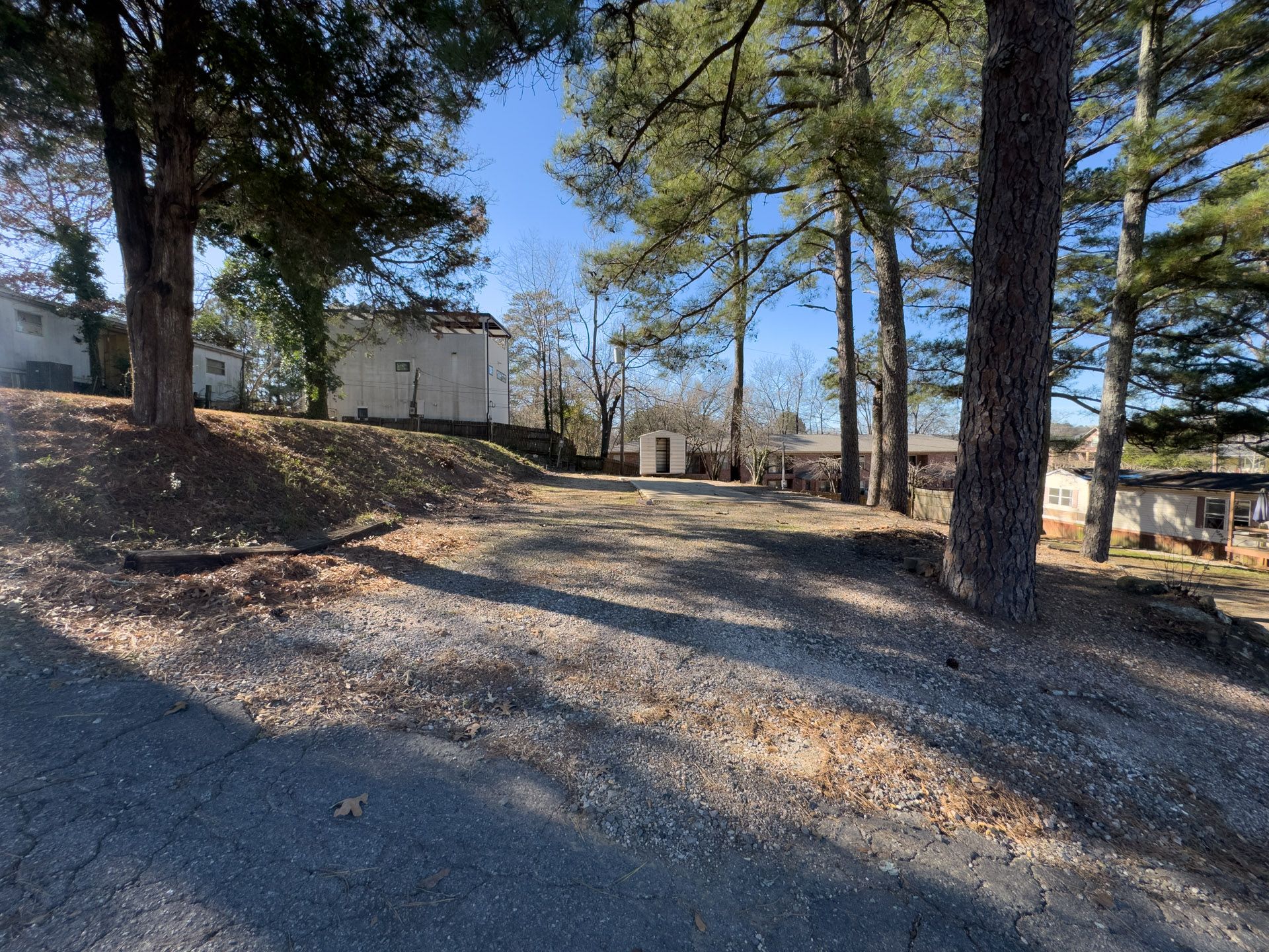 A gravel driveway leads towards several low buildings under a blue sky, trees on both sides.