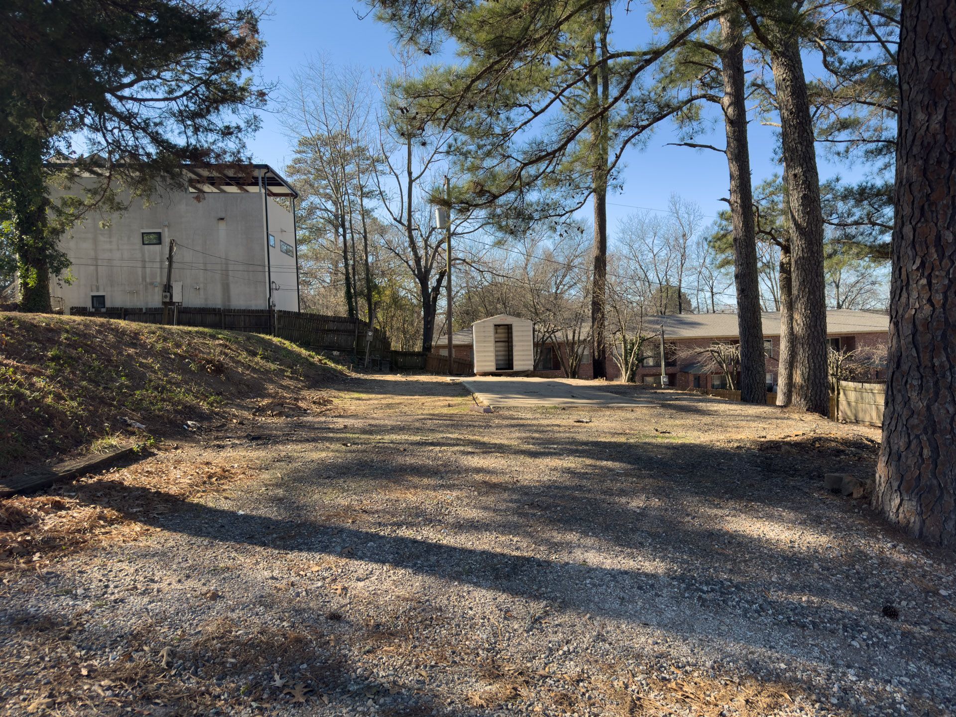 Gravel driveway leading to structures under a clear sky. Trees frame the view, casting long shadows.