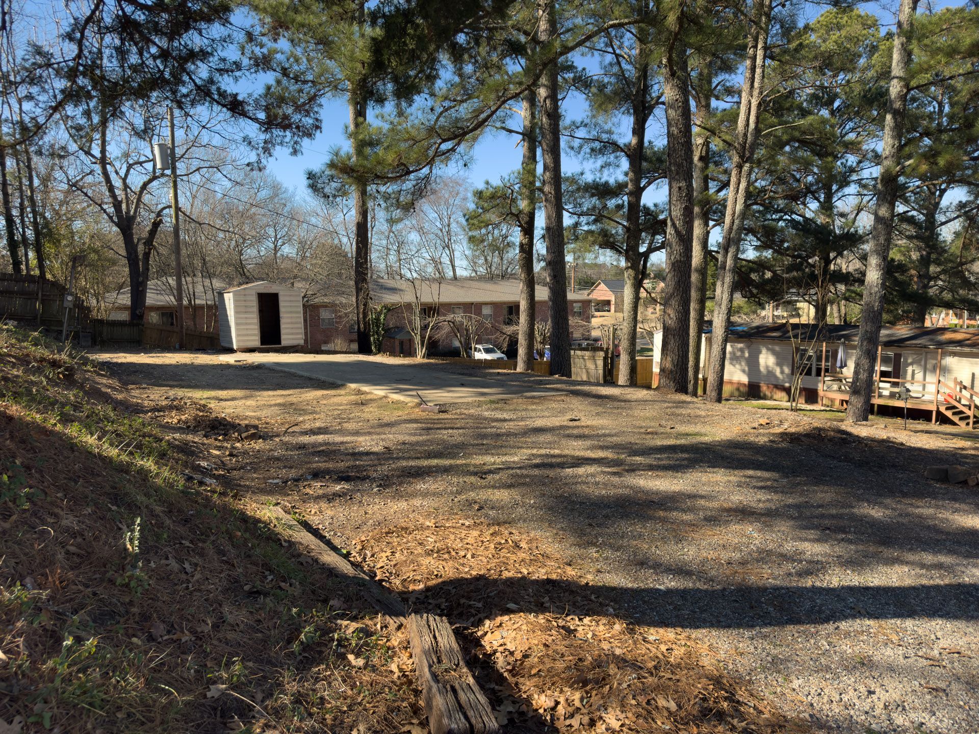 Gravel lot with shed, trees, and buildings in the background under a blue sky.