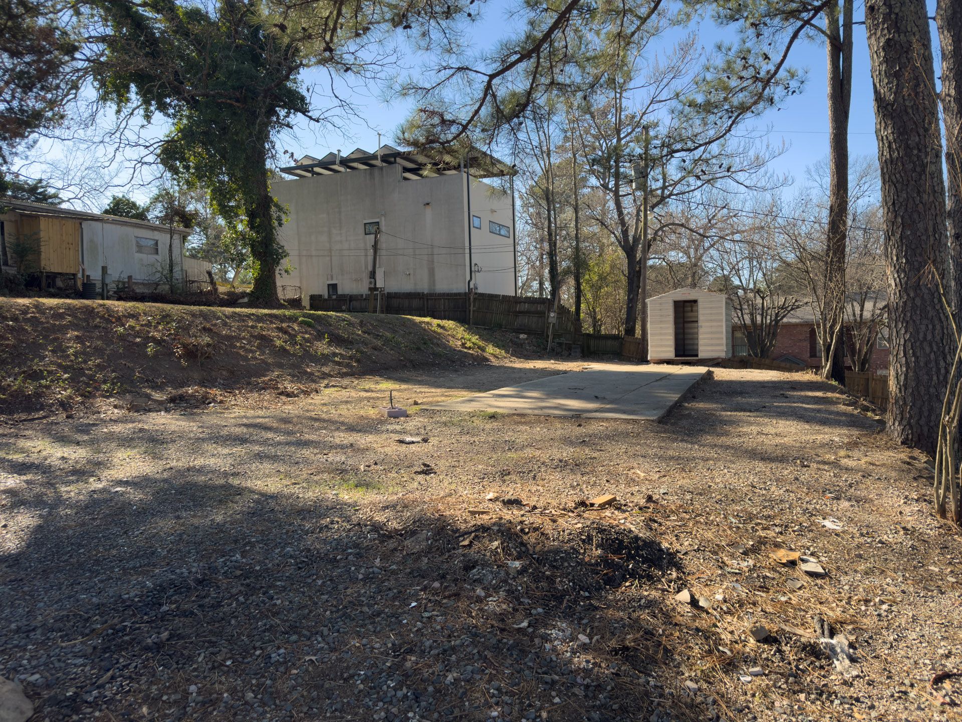 Gravel lot with shed, two-story white building, and trees on a sunny day.