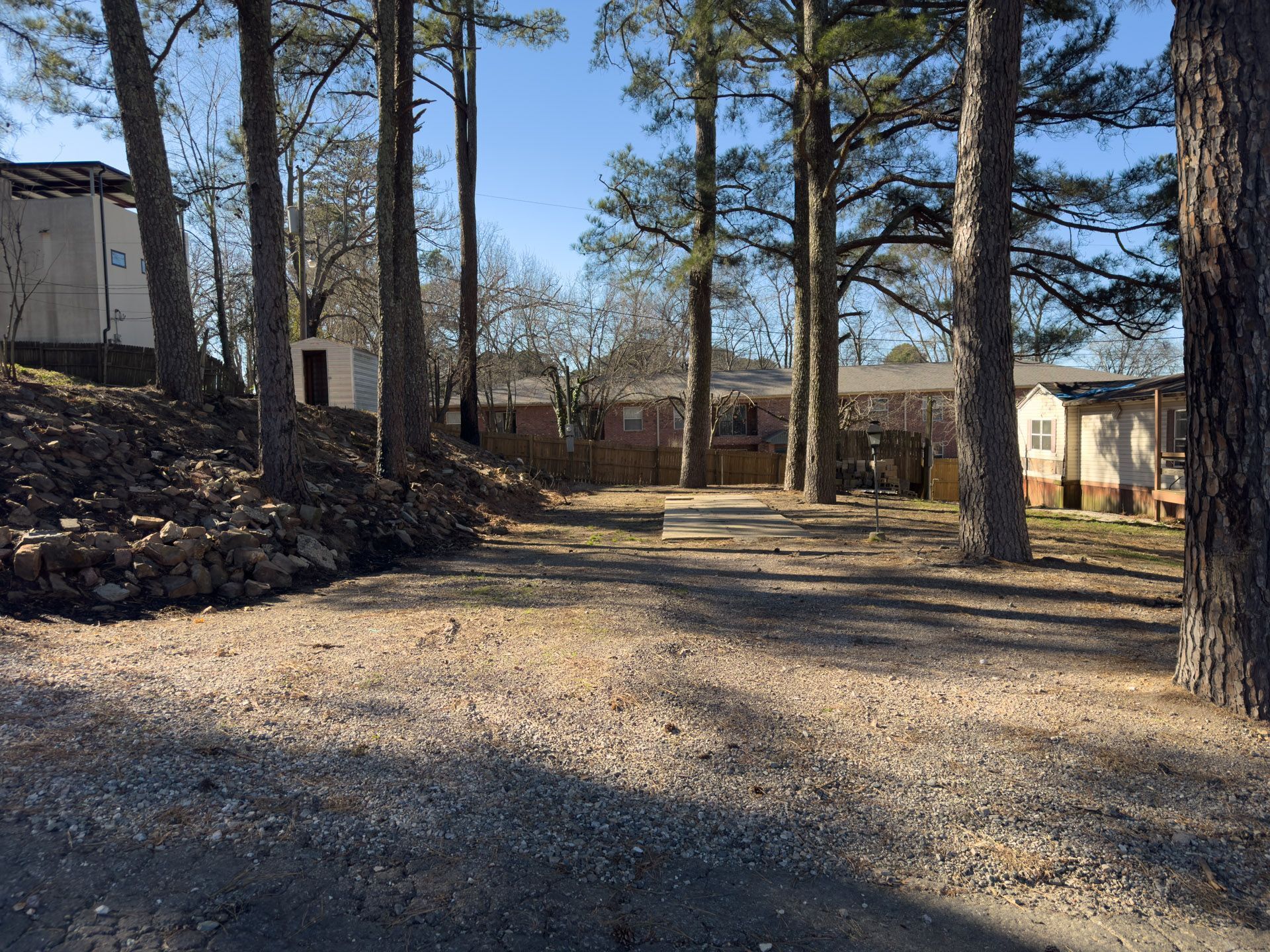 Gravel driveway surrounded by trees, leading to a yard with a concrete ring and houses in the background.