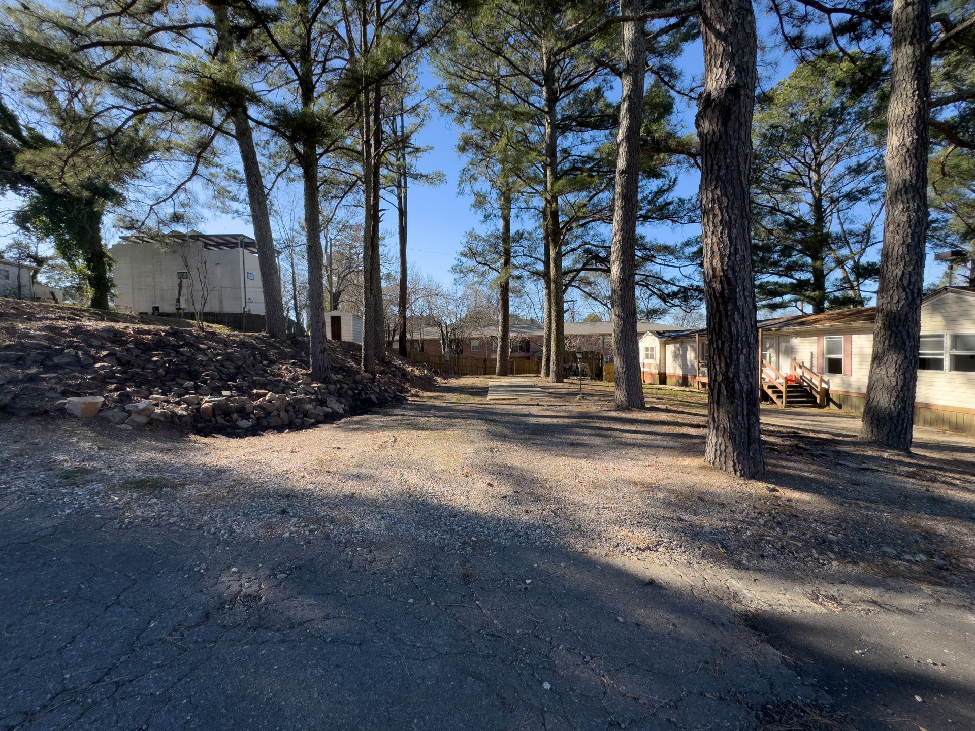 Gravel road leads through trees toward low buildings under a blue sky.