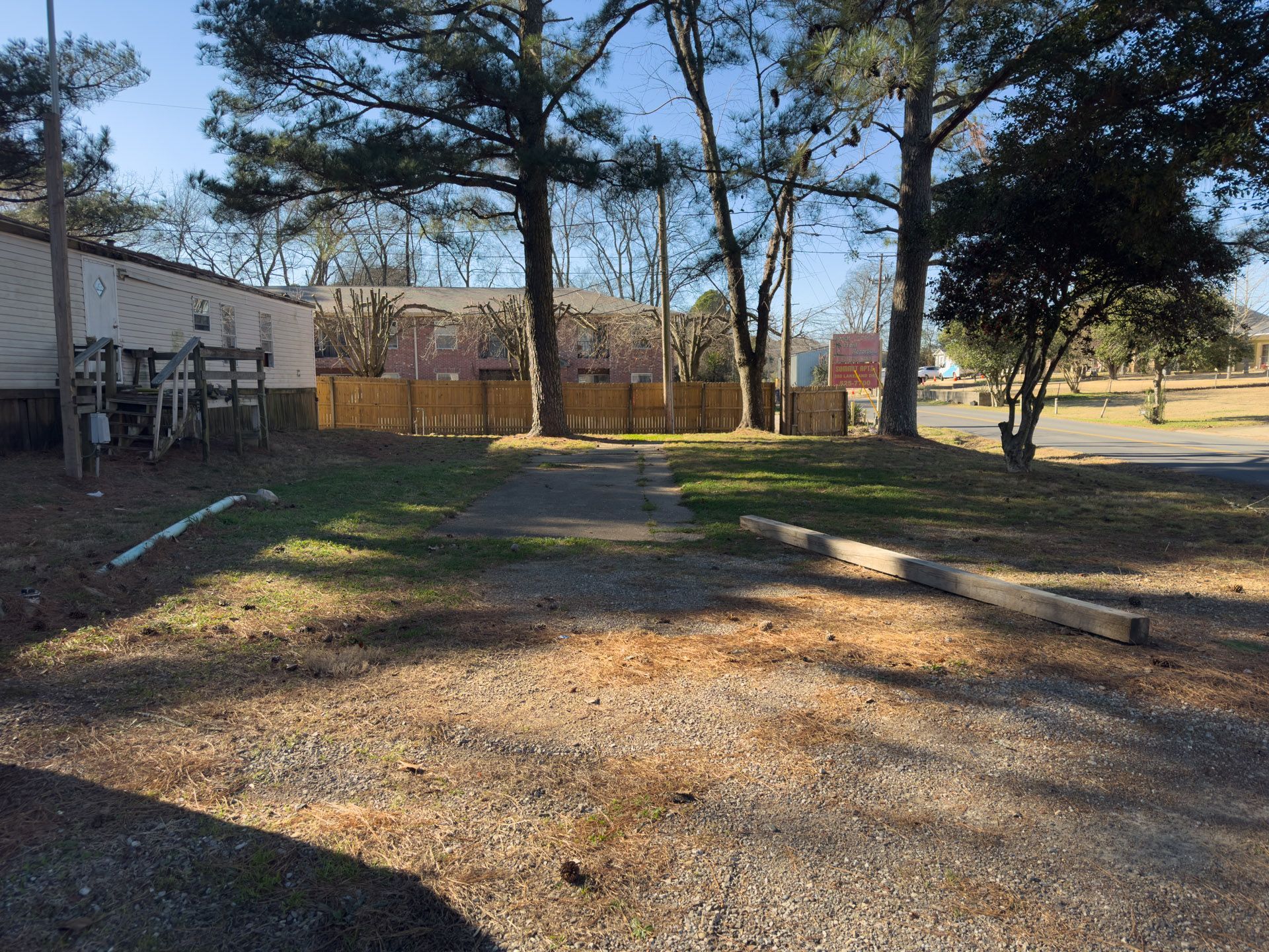 Gravel parking area leads to a grassy space with a pathway. A wooden fence and trees are visible in the background.