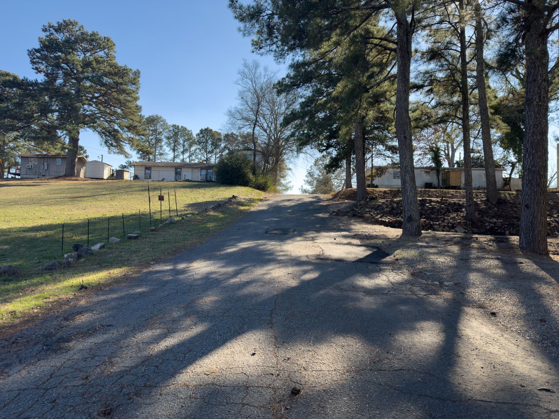 Gravel road leading uphill, flanked by trees and a mobile home on each side, under a clear sky.