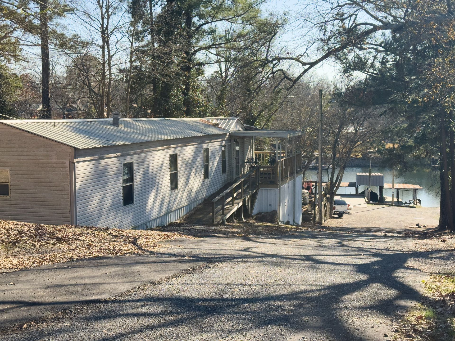 Mobile home with sloped roof, wooden porch, and steps leading to a gravel driveway. Trees and water in the background.