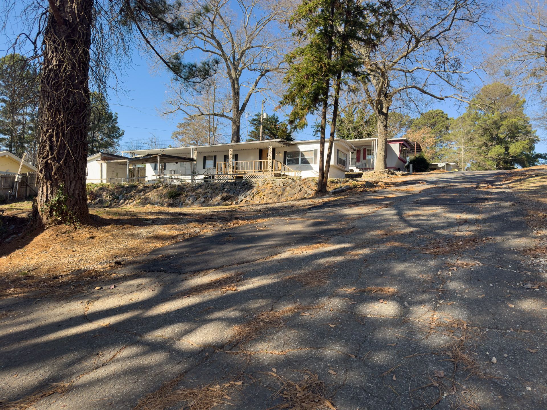 A gravel driveway leads to a long, one-story house with a porch, trees, and a clear blue sky.