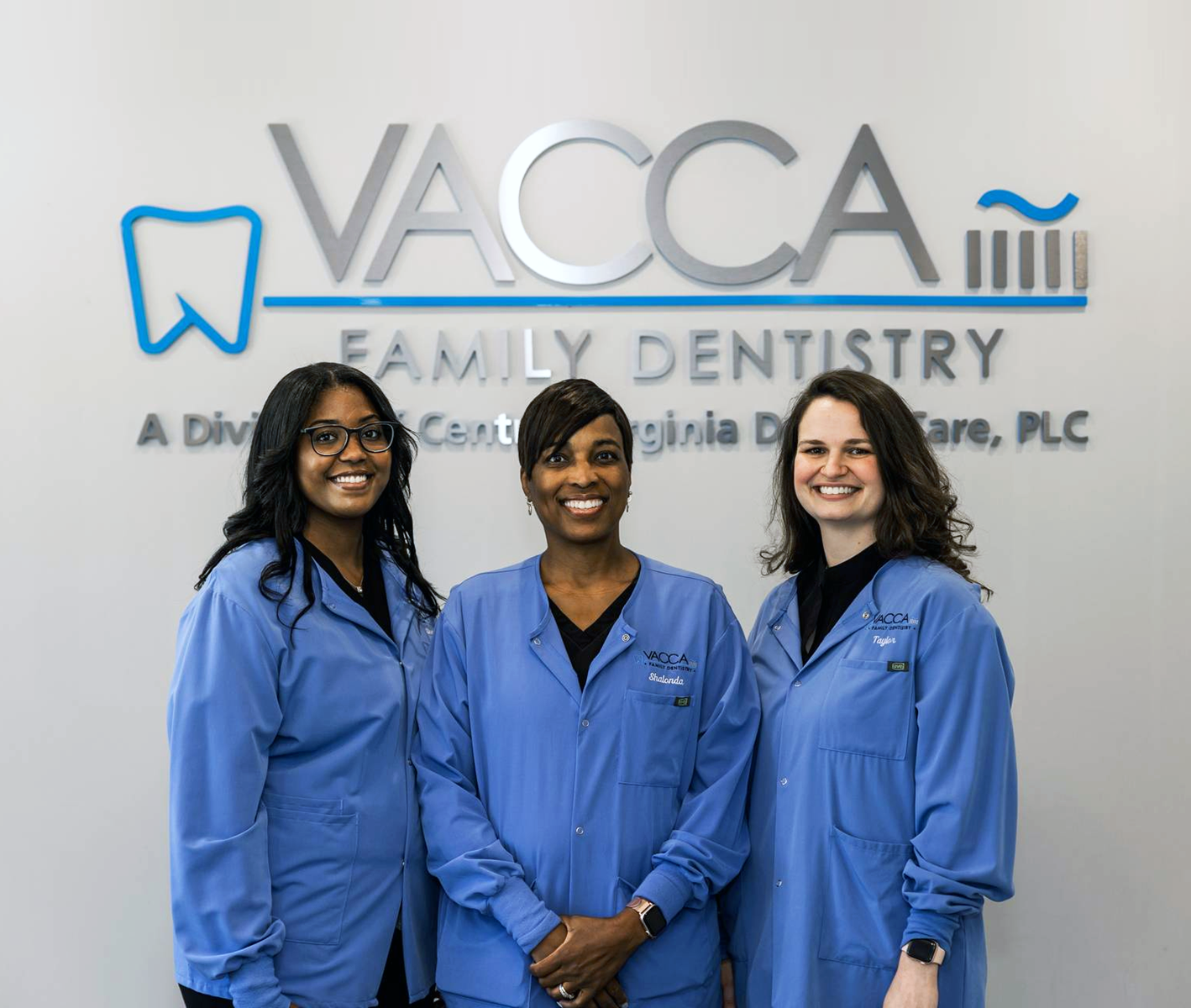 Three women are posing for a picture in front of a vacca family dentistry sign.
