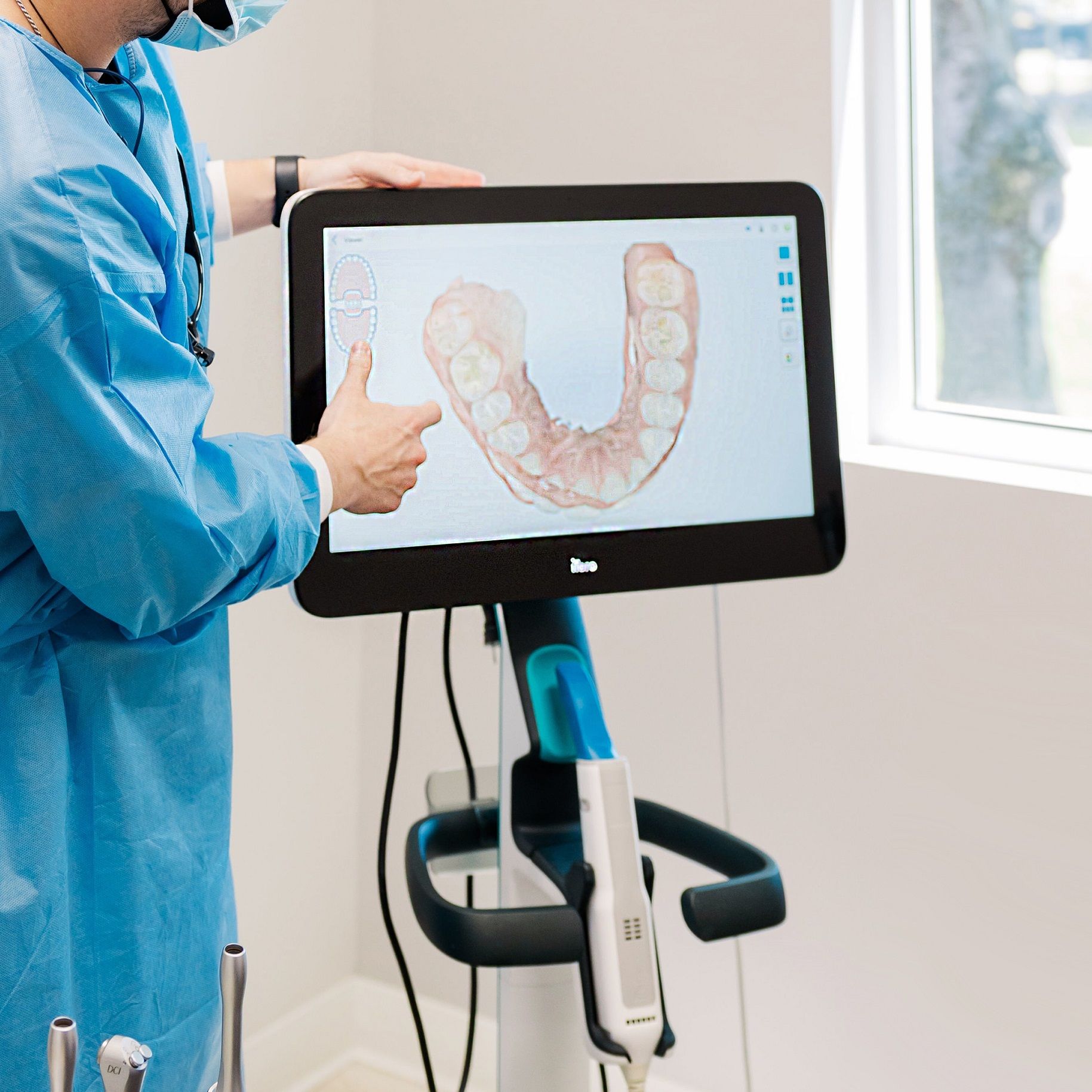 A man in a blue gown is pointing at a monitor with a picture of teeth on it
