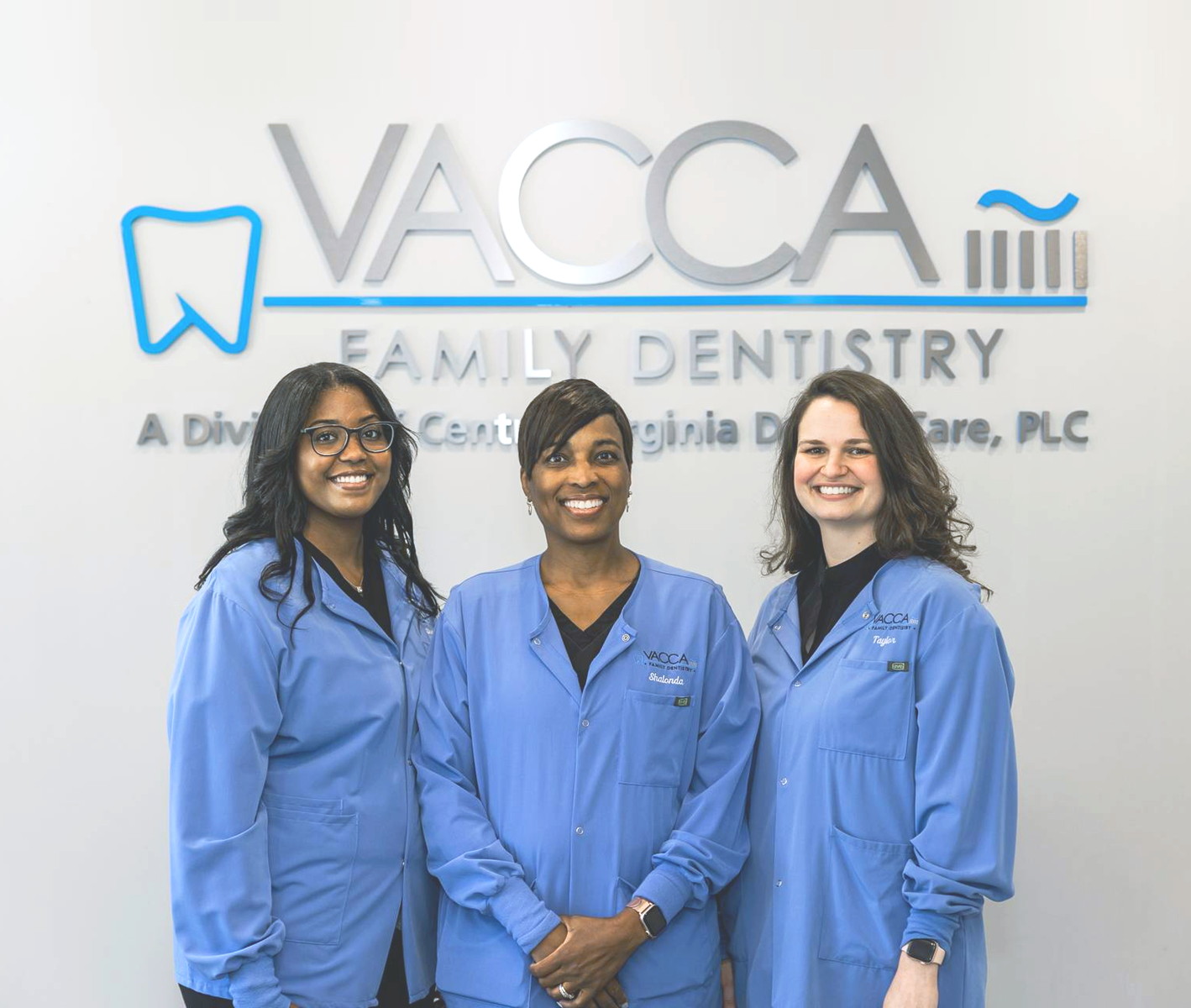Three smiling dental staff in blue coats stand in front of a Vacca Family Dentistry sign.