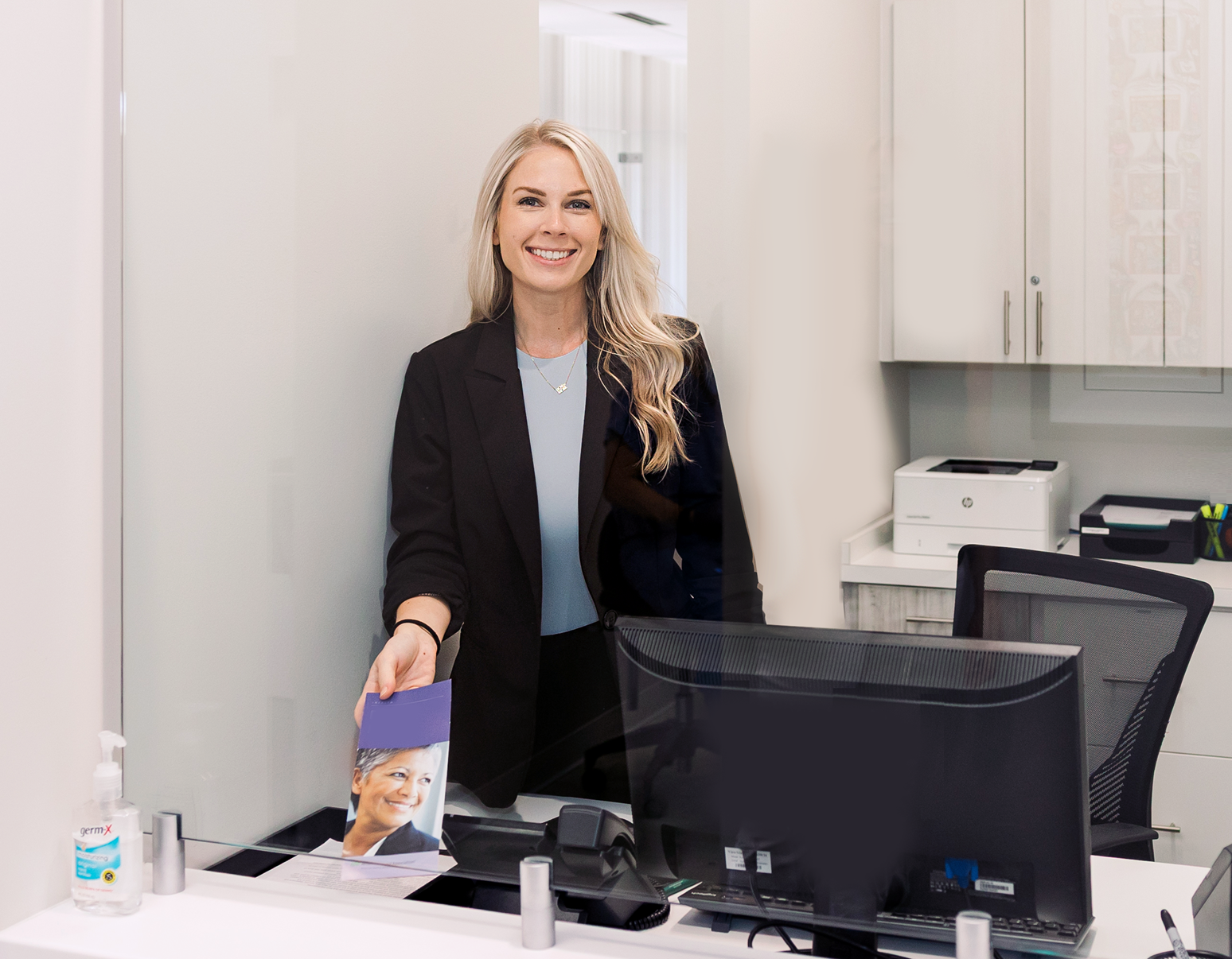 Woman standing behind a desk in an office, holding a brochure beside a computer monitor.