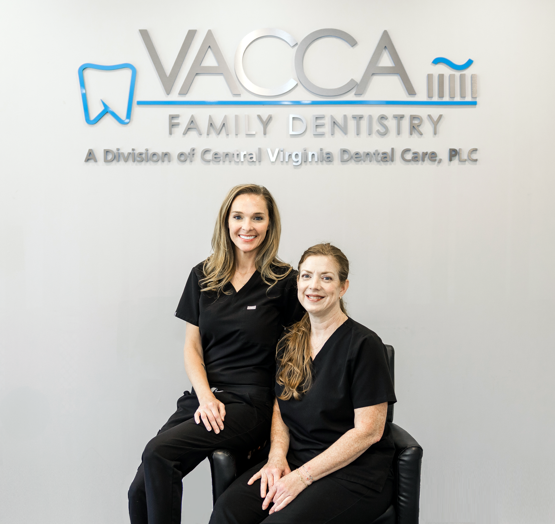Two women are posing for a picture in front of a sign for vacca family dentistry.