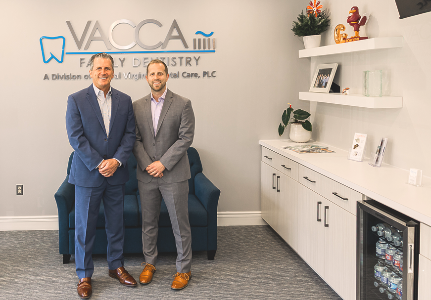Two men in business suits standing in a bright office lobby with VACCA Family Dentistry signage