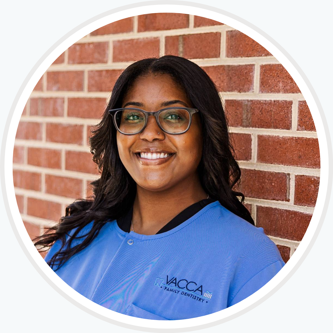 A woman wearing glasses and a blue shirt is smiling in front of a brick wall.