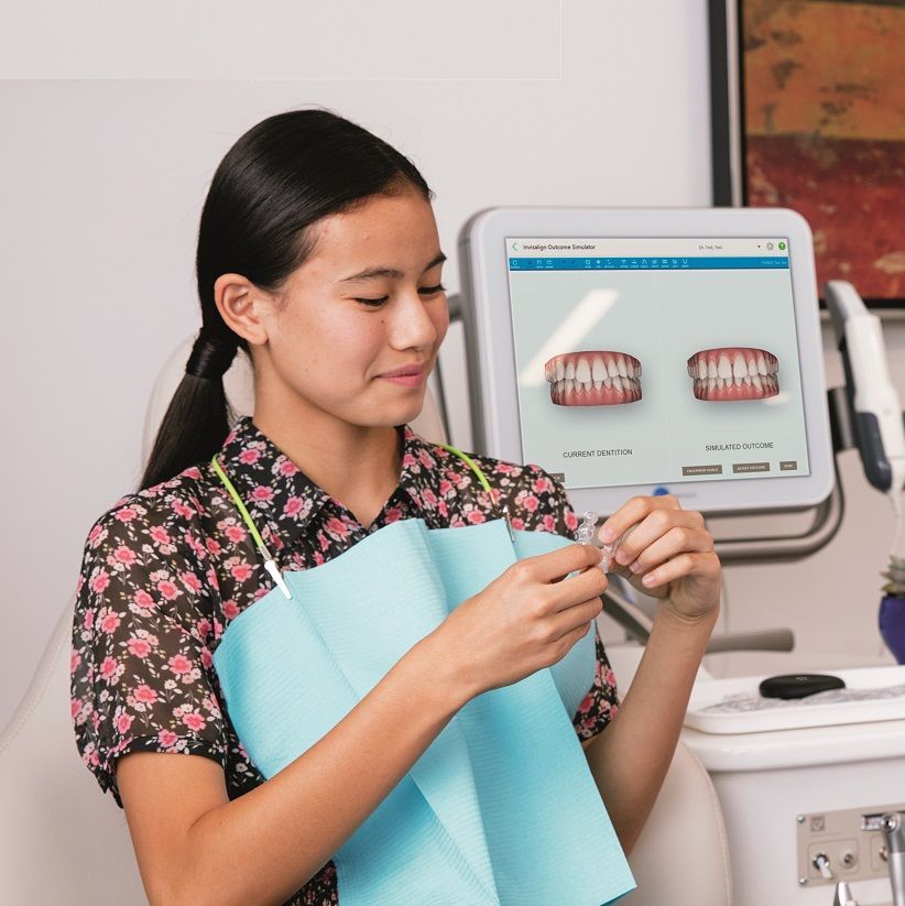 A woman in a floral shirt is holding a blue apron in front of a monitor showing teeth