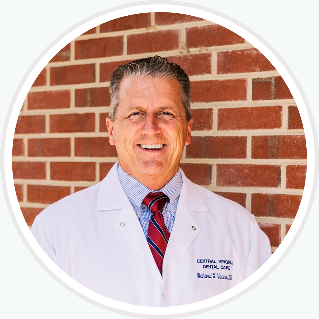 A man in a lab coat and tie is smiling in front of a brick wall