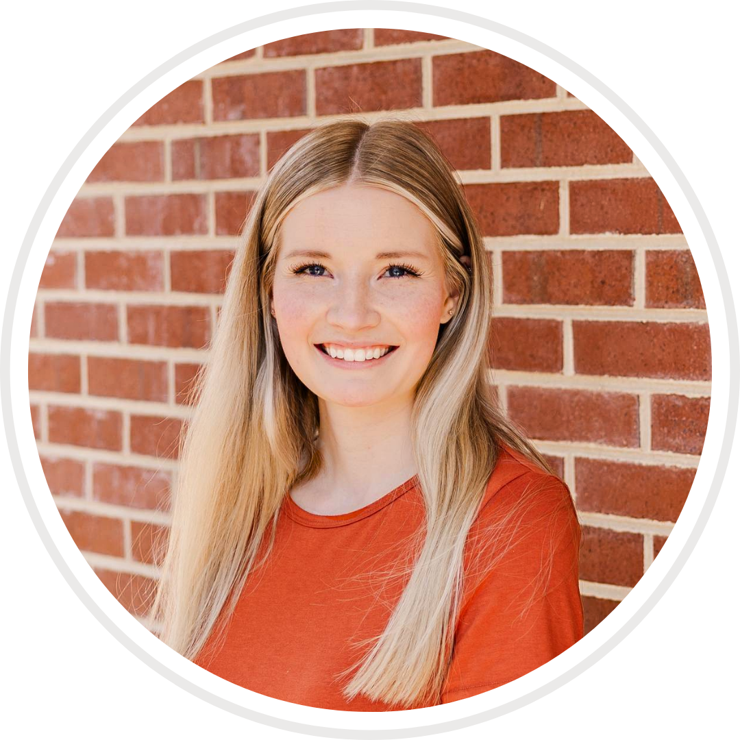 A woman in an orange shirt is smiling in front of a brick wall.