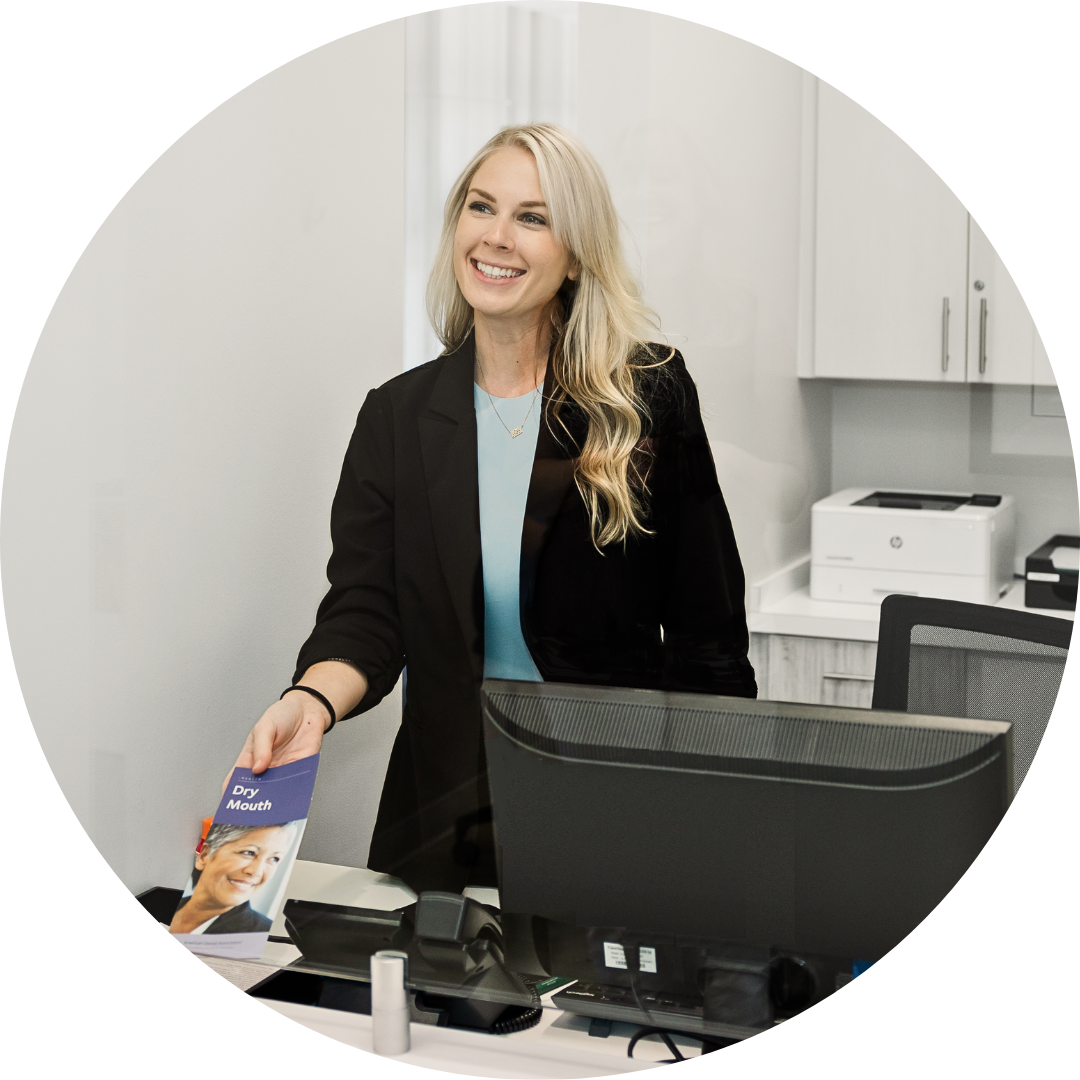 A woman is standing at a desk in front of a computer and smiling.