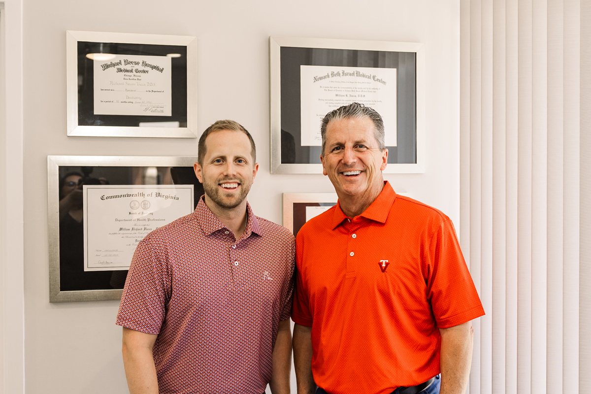 Two men standing indoors beside framed certificates, one in a patterned shirt and one in an orange polo.