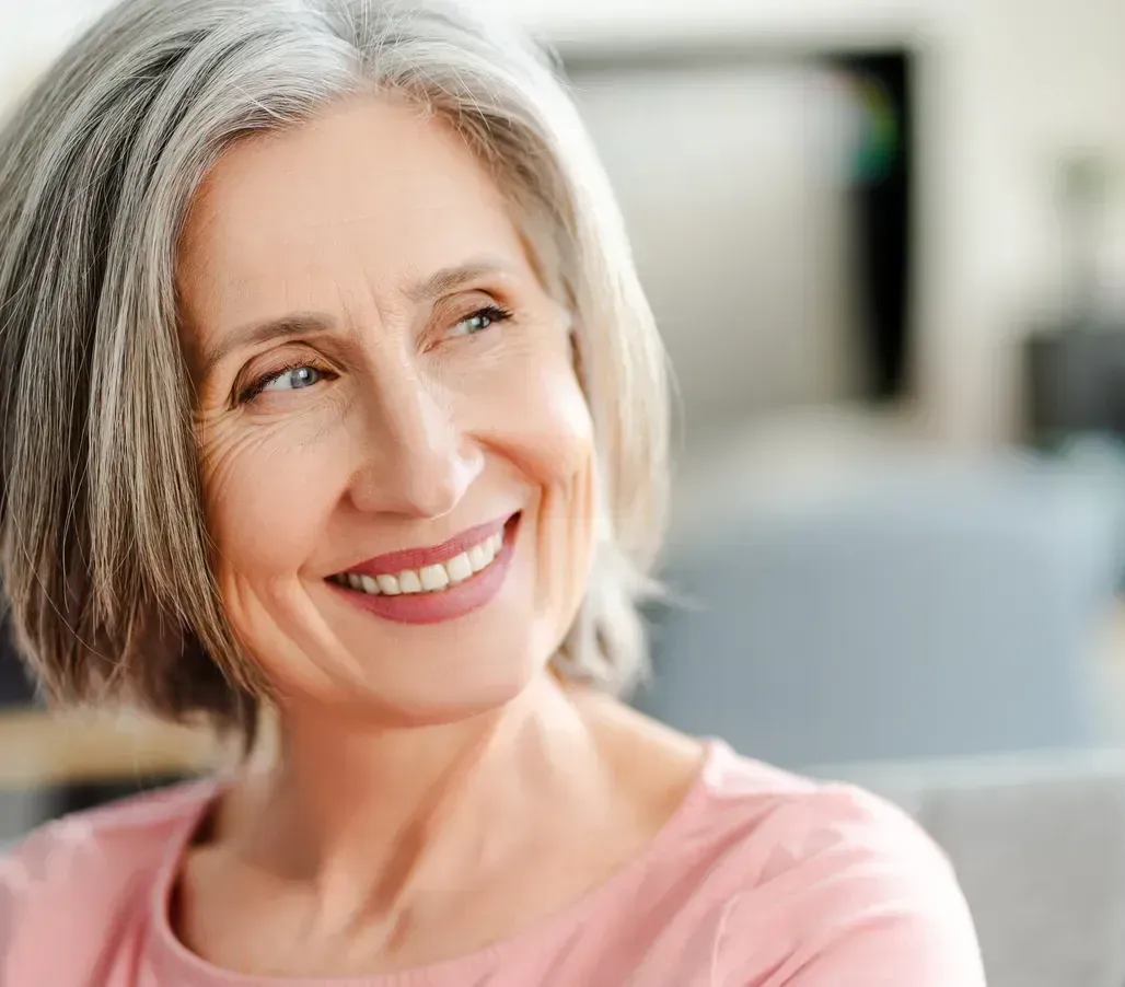 Smiling older woman with gray hair looking off to the side; wearing a pink top, seated indoors.