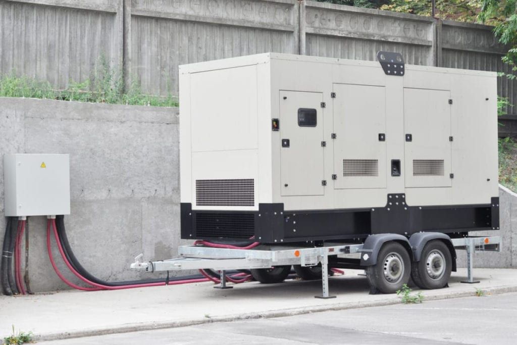 Large beige generator on a trailer parked beside a gray wall, connected to an electrical box.
