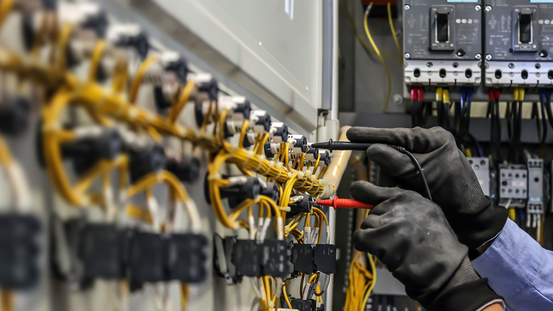 Electrician using a multimeter to test wires in an electrical panel, wearing black gloves.